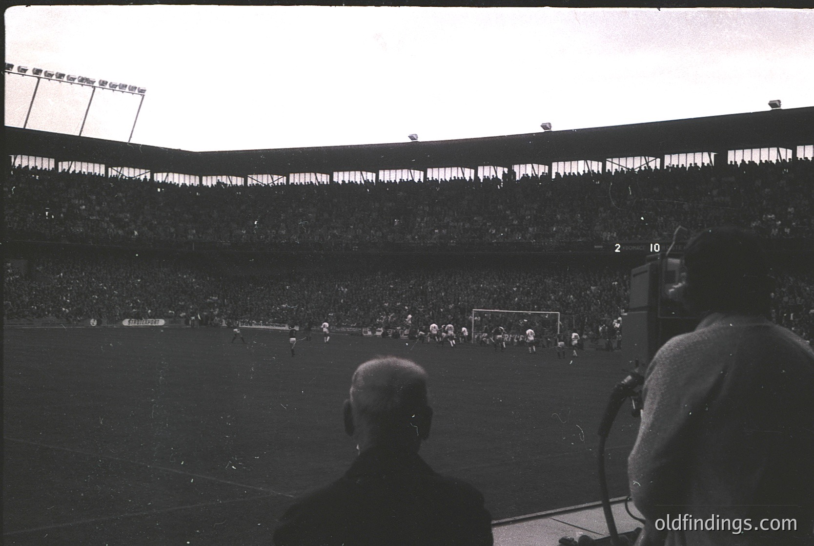Vintage black-and-white shot of a packed stadium during a soccer match, likely from the 1950s–1960s. Crowds fill tiered seating, with players and a referee visible on the field. Overhead floodlights and scoreboard indicate evening play. Spectator’s back (with microphone) suggests broadcast or commentary.