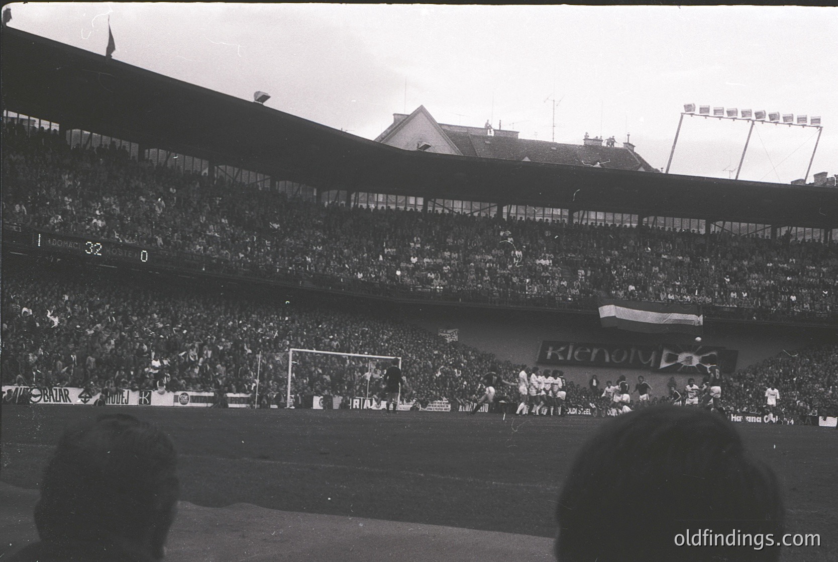 Vintage black-and-white stadium shot showing packed stands with dense crowds, likely during a 1960s–1970s European football match. Visible scoreboard reads "32-0" and "KENOLYBUS" branding. Players in white uniforms near the field; architectural elements include tiered seating and floodlights. Crowd density and banners suggest high-energy match atmosphere.