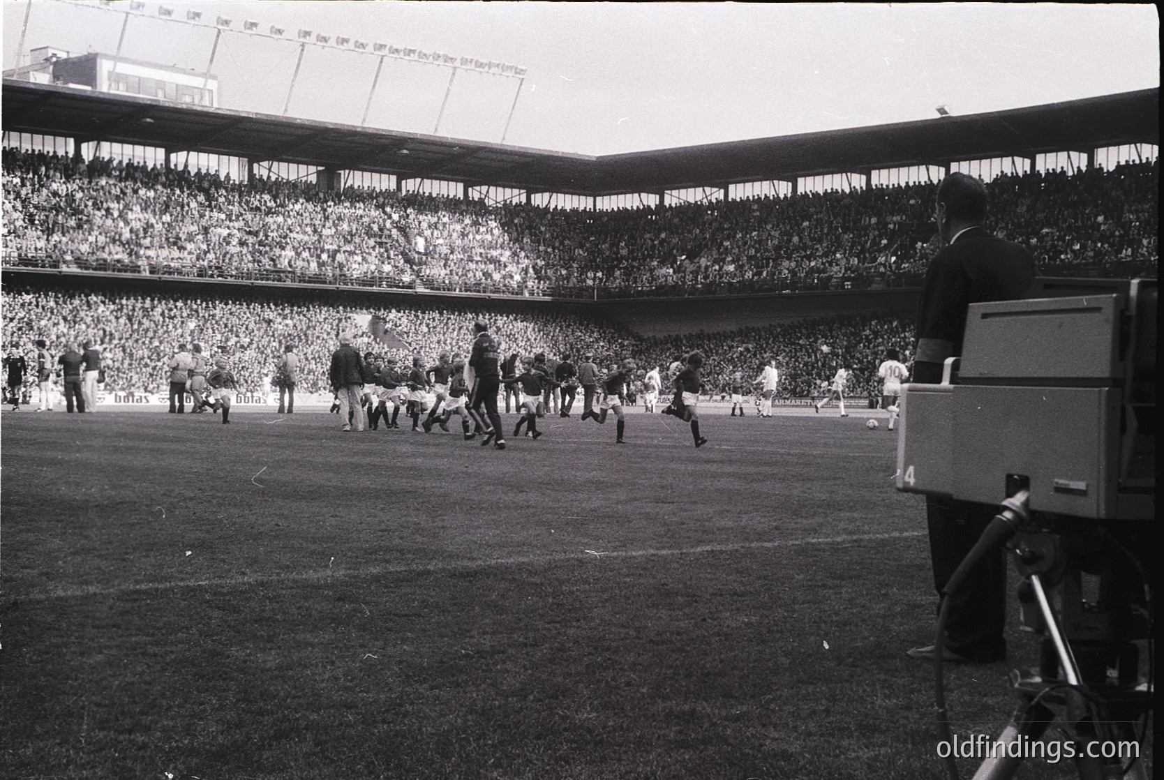 Vintage black-and-white shot of a packed stadium during a mid-20th-century football match. Players in shorts and boots engage in action on the field, while a broadcast camera crew captures the event. Tiered seating filled with spectators, some waving scarves. Architectural details include curved stands and a visible stadium sign.