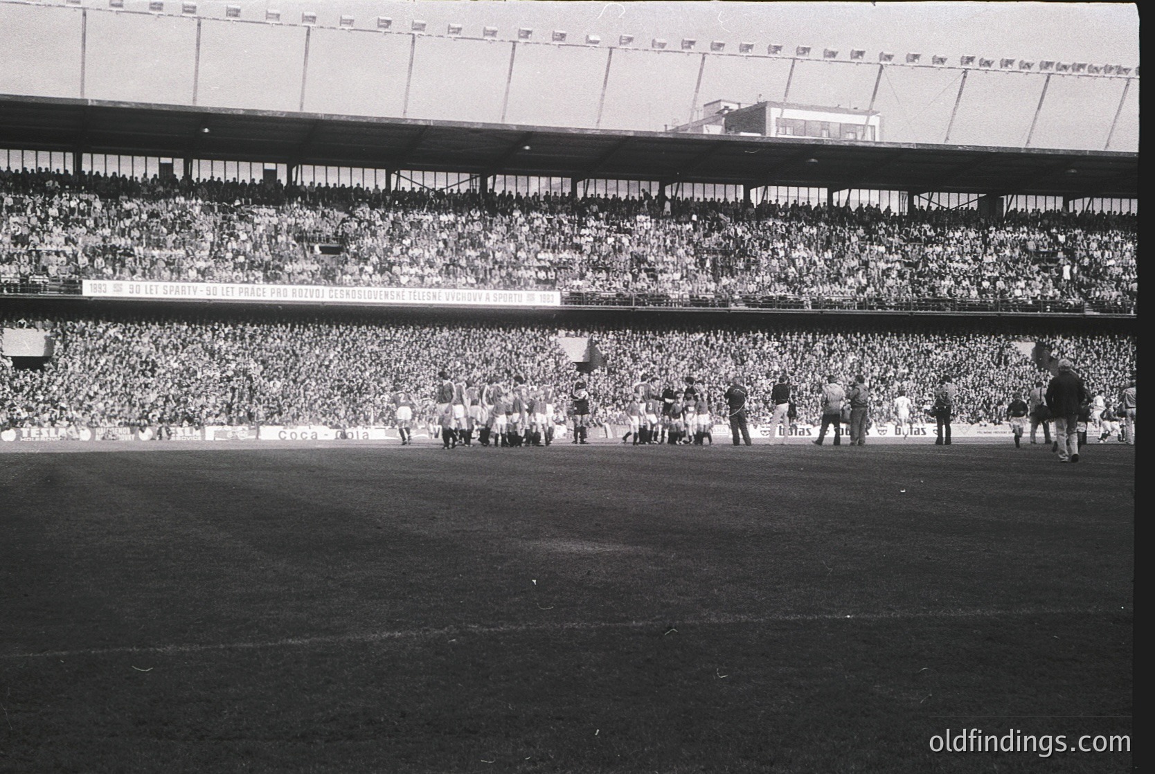 Vintage black-and-white stadium shot showing packed stands with dense crowds, likely UEFA/European Cup final. Players in white kits advance toward goal, flanked by officials. Mid-20th century European football atmosphere.