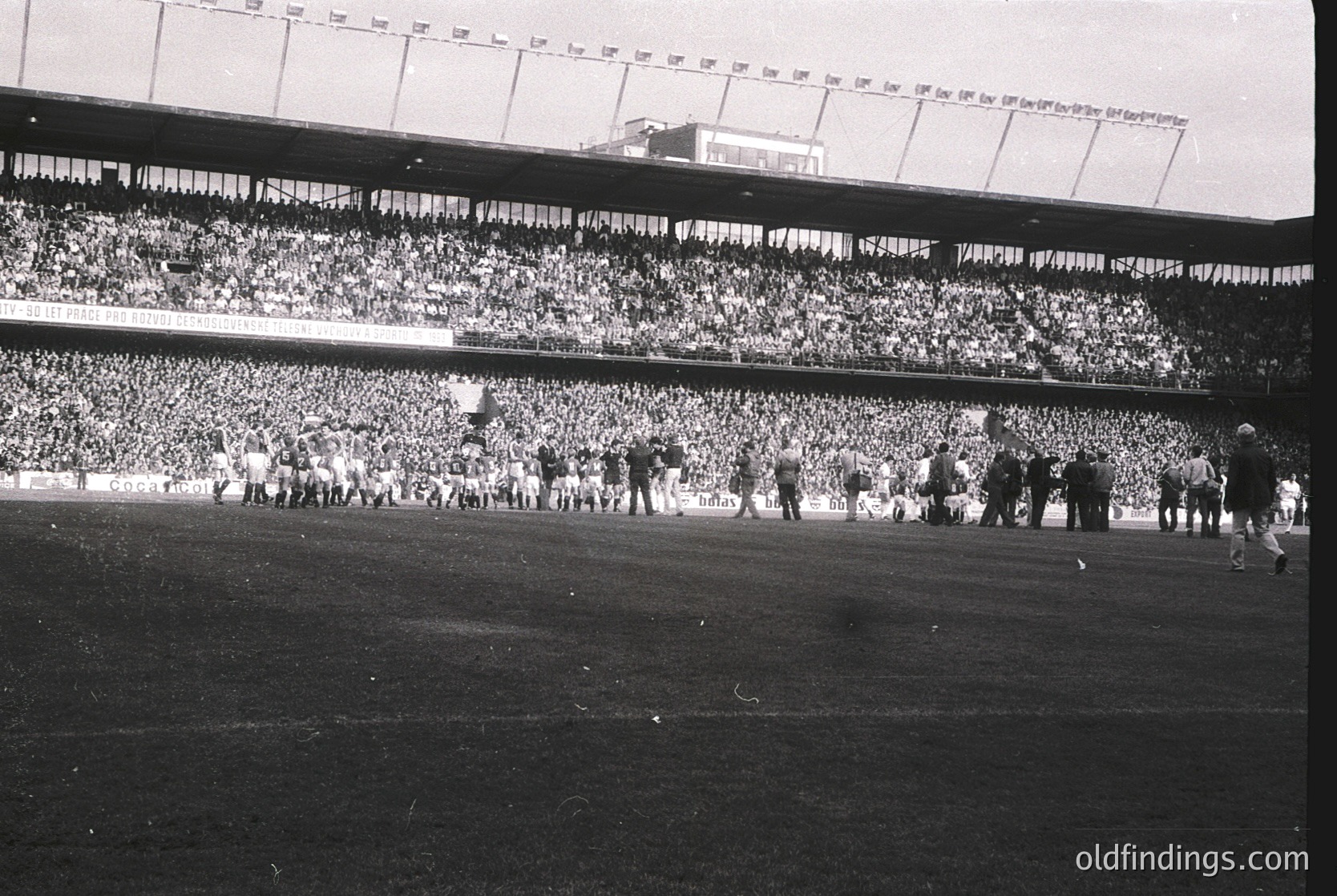 Vintage black-and-white stadium shot showing packed tiered seating with dense crowds, likely a 1950s–1960s European football match. Players in white uniforms advance toward the camera, while spectators fill all levels. Advertising banners (e.g., "CP") and stadium signage visible. Ideal for sports history, retro design, or cultural studies.