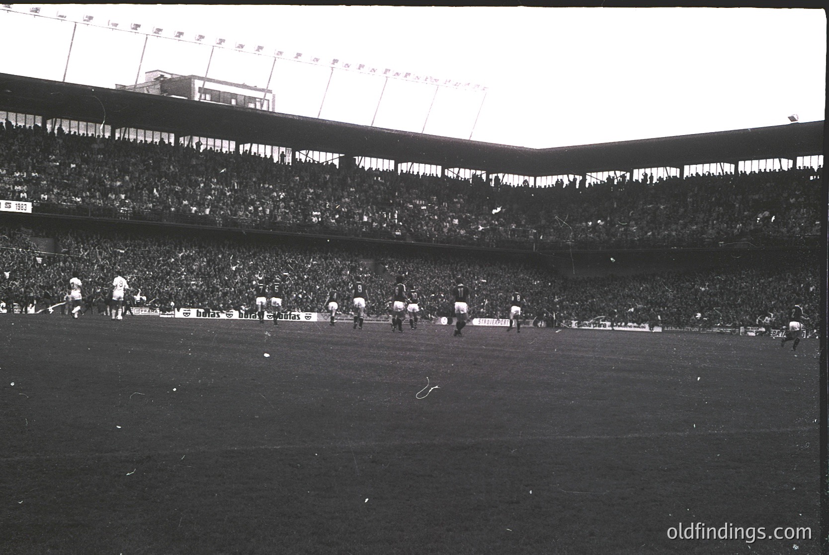 Vintage black-and-white shot of a packed stadium, likely from the 1950s–1960s. Massive crowd fills tiered seating under floodlights, with players in white uniforms mid-match on a grass field. Architectural details include curved stands and exposed stadium infrastructure.