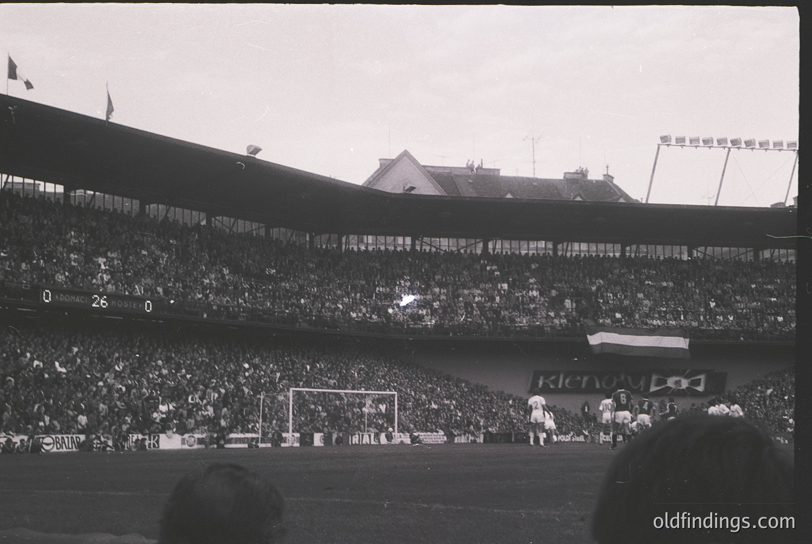 Vintage black-and-white stadium shot showing packed stands with dense crowd. Players in white uniforms near goalpost; visible scoreboard reads "26-0." Advertisement for "Klenov" on field. Architectural elements include tiered seating and floodlights. Likely Eastern Bloc-era match, 1960s-70s.