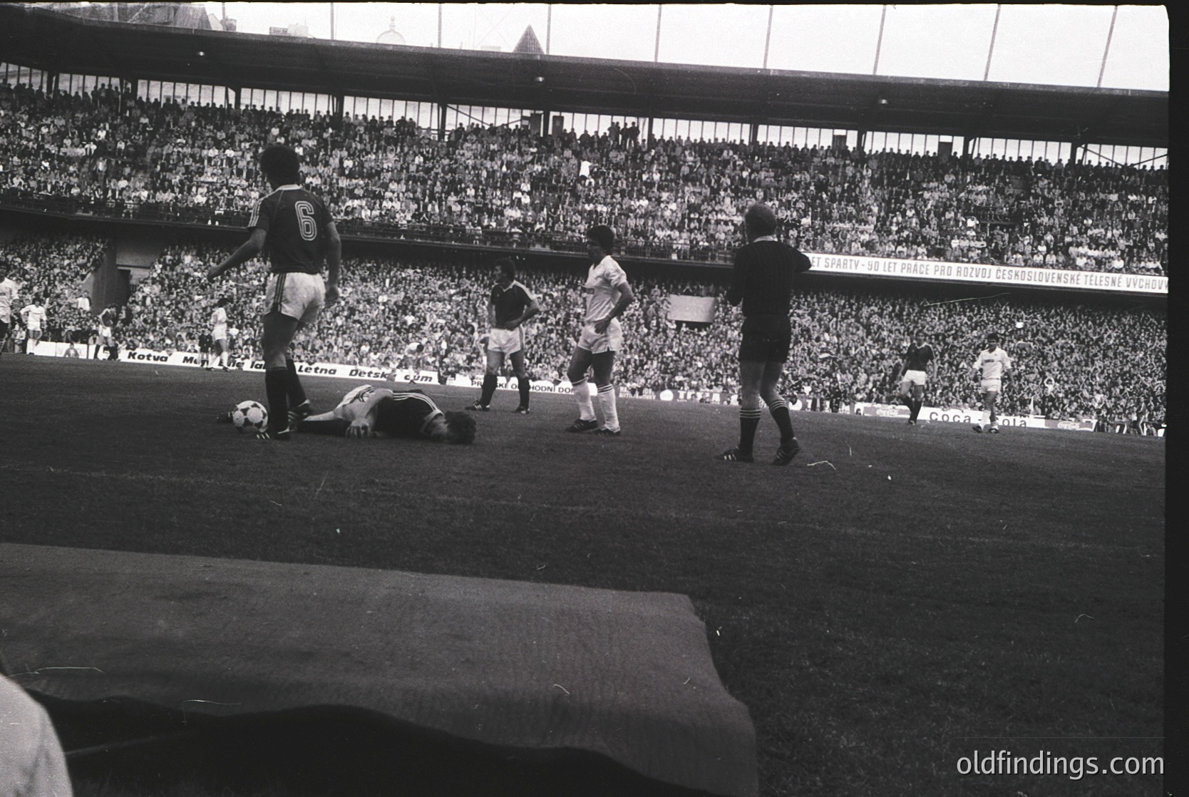 Black-and-white shot of a 1970s soccer match in a packed stadium, featuring a fallen player and teammates reacting. Uniforms suggest European league play, with visible sponsor logos on jerseys. Crowd fills stands, indicating high attendance.
