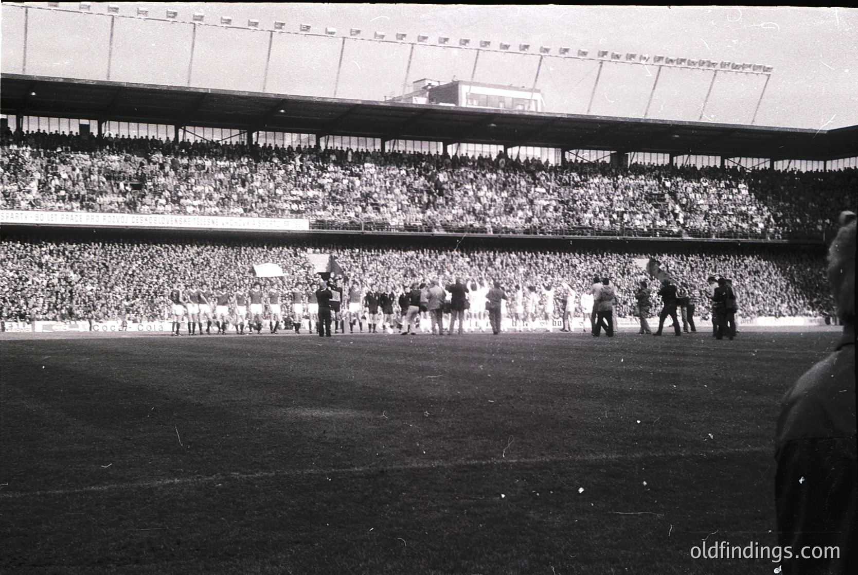 Vintage black-and-white stadium shot showing packed tiered seating with dense crowds. Players in vintage uniforms line up on the field, likely pre-game or halftime. Architectural details include tiered seating and stadium signage. Mid-20th century European football atmosphere.
