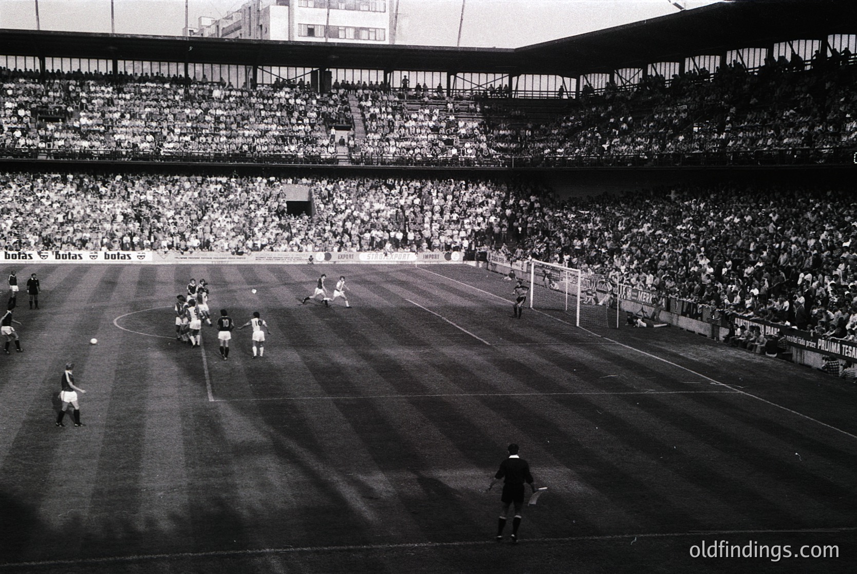 Classic black-and-white shot of a packed stadium during a mid-20th century soccer match. Players in vintage striped jerseys engage in action near the goal, with a referee overseeing. Crowd fills tiered stands, creating a sea of spectators. Signs in Spanish ("butas" and "huevos") hint at a lively, possibly Spanish-speaking fanbase. Architectural details suggest a large, historic venue.