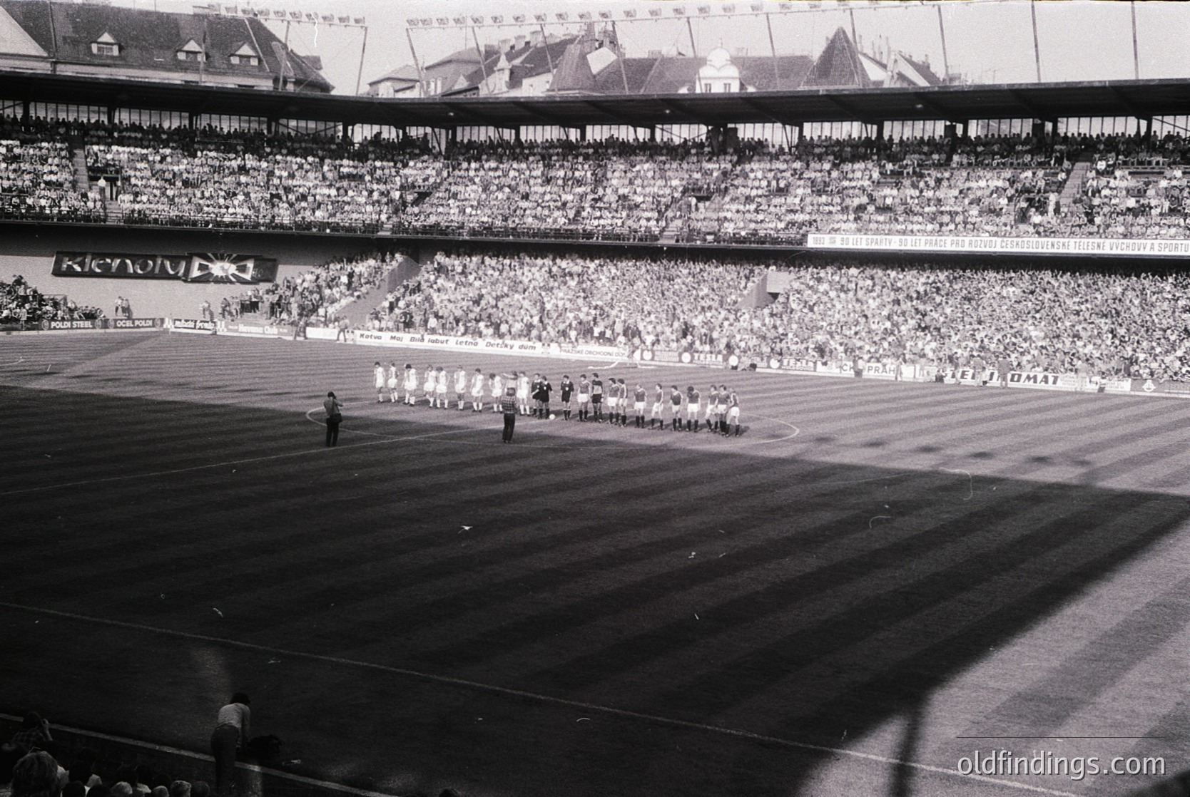 Vintage stadium shot featuring a large crowd in tiered seating, likely from the 1950s–1960s. Central field shows a marching band or ceremonial procession in formation. Architectural details include curved stands and visible stadium branding (). Crowd density and attire suggest a major public event or sports occasion.