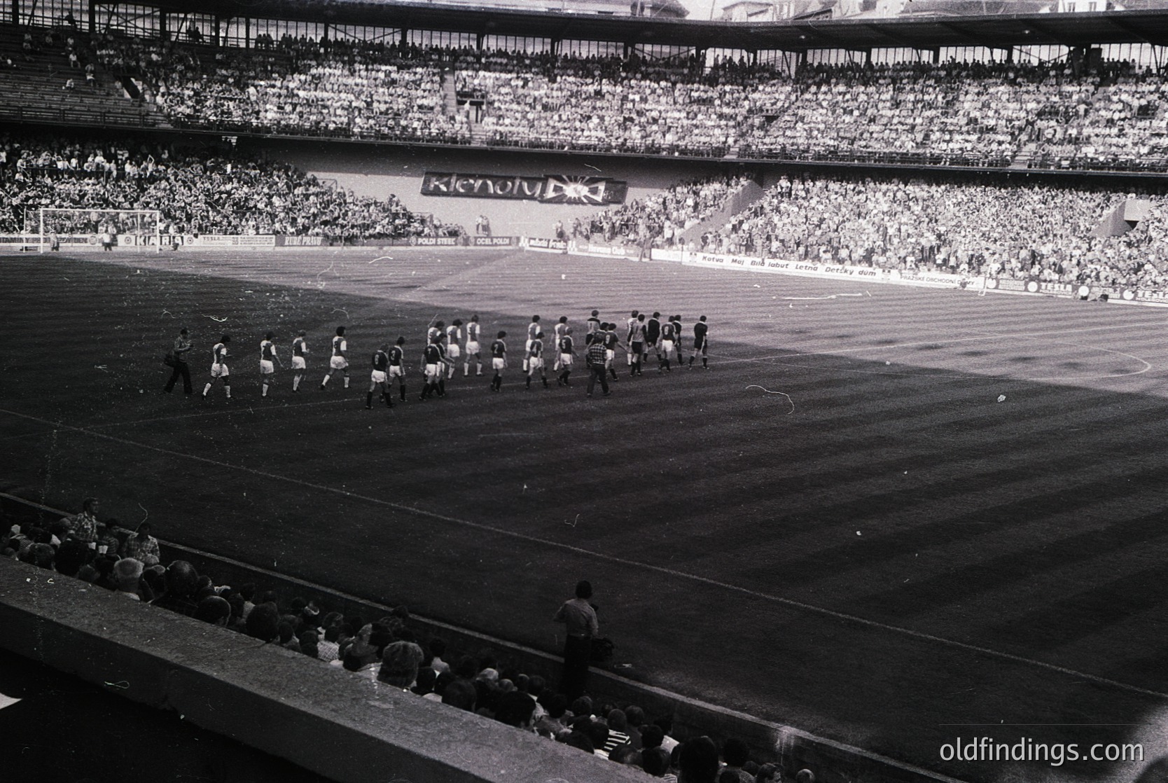 Black-and-white stadium shot showing a pre-game ceremony with teams lined up on the field, likely mid-20th century. Massive crowd fills tiered stands; banners and flags suggest national or club affiliation. Distinctive tiered seating and stadium architecture hint at European design.