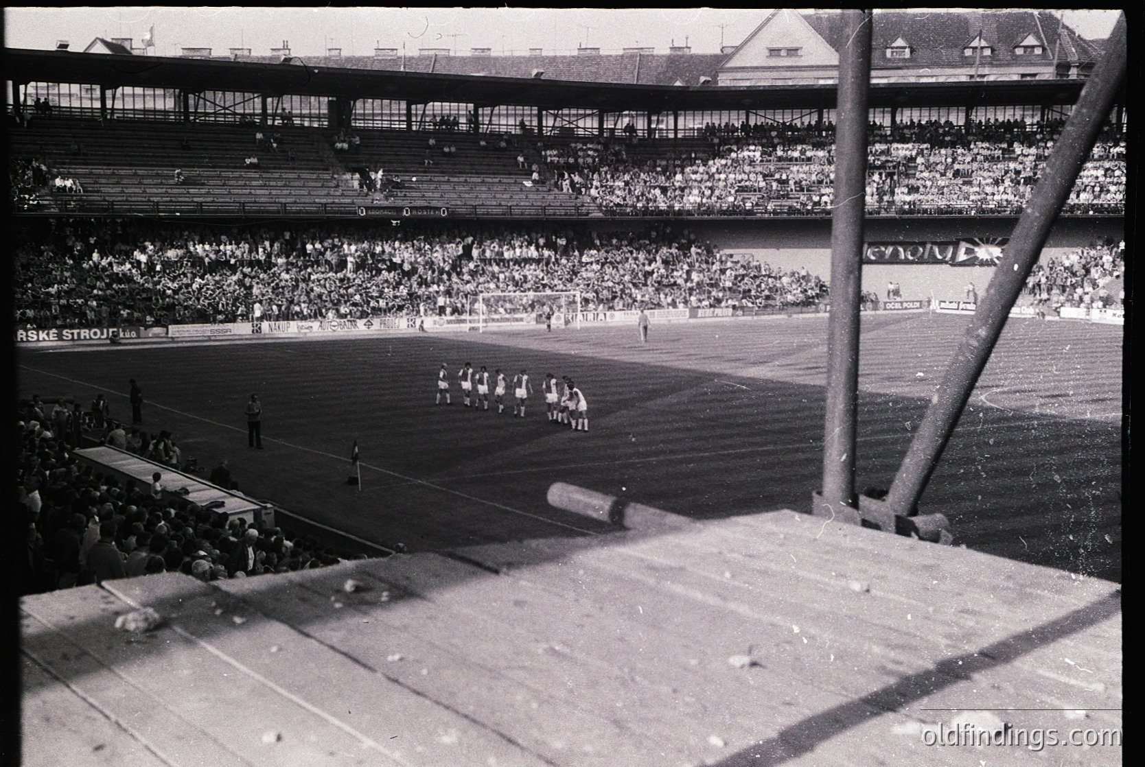 Mid-20th century stadium scene with packed stands, likely European. Players in white uniforms on a grass field, surrounded by dense crowds. Advertisement boards display "ASKE STROJ" and "KONFERENZ." Architectural details include tiered seating and exposed concrete supports.