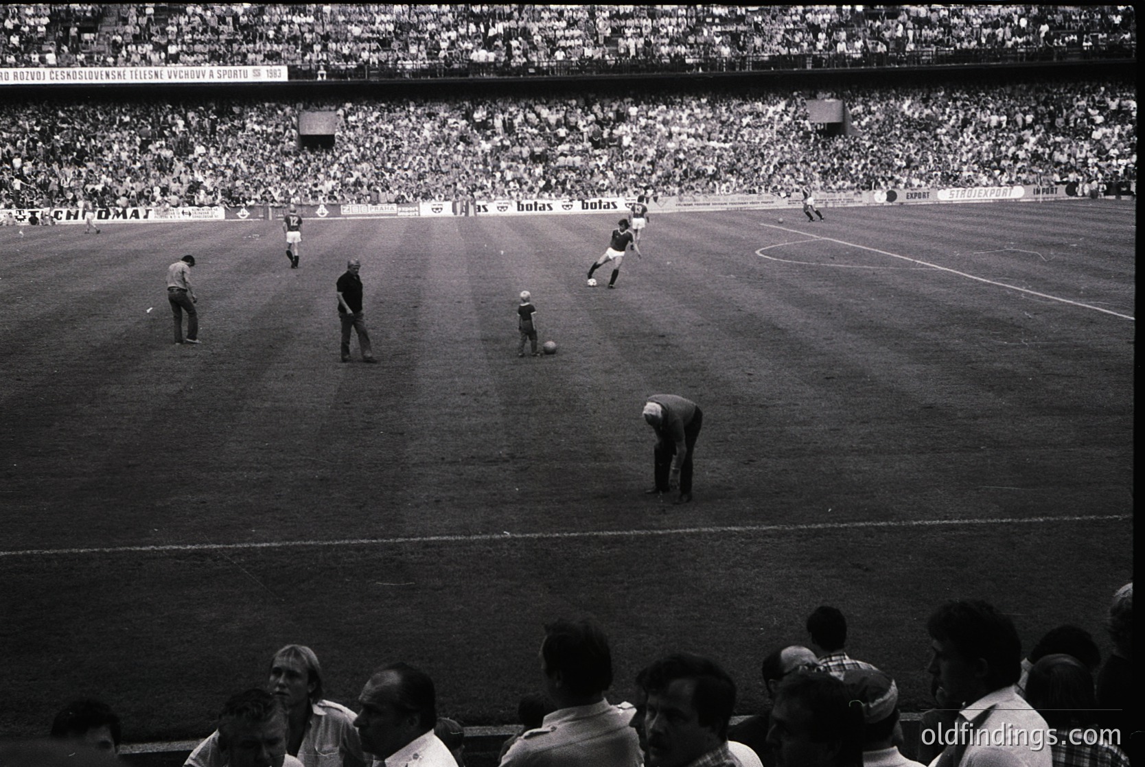 Vintage black-and-white soccer match in a packed stadium, featuring midfield action with players in classic striped jerseys. Referees and linesmen oversee play on a grass pitch. Crowd fills stands, suggesting 1960s–1970s European football culture. Ideal for sports history, nostalgia, or retro design references.