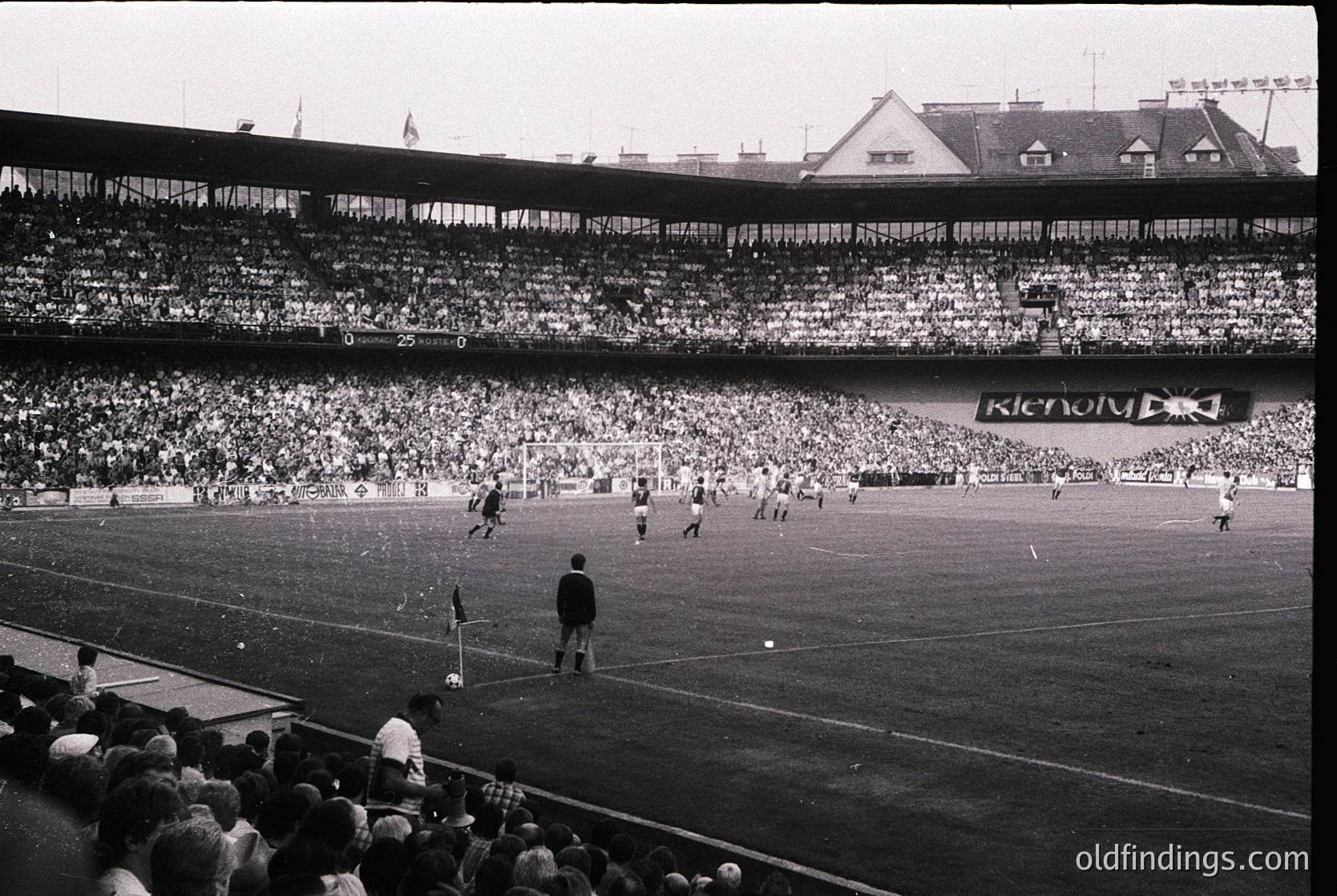 Vintage black-and-white stadium shot showing a packed 1960s-era football match. Multi-tiered seating filled with spectators, visible scoreboard displaying "0-0," and "Kenody" advertisement on the stand. Grass pitch with players in action and a referee overseeing play. Architectural details include curved roofing and tiered seating.