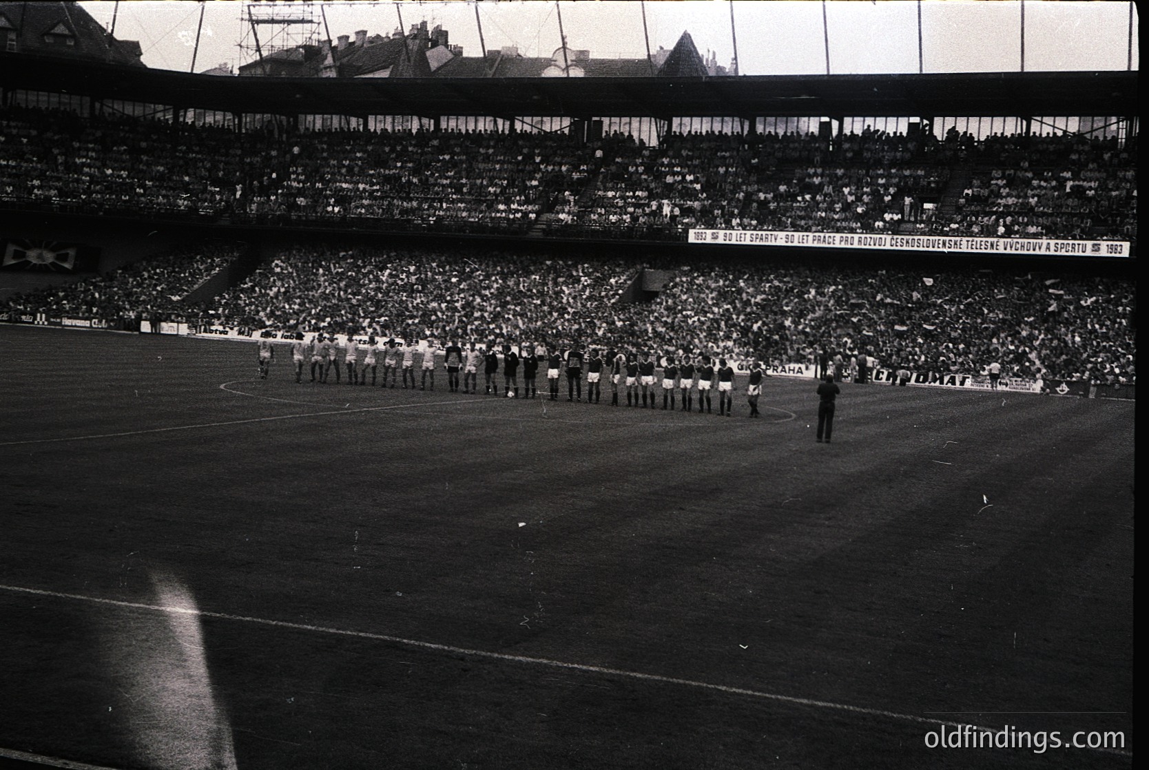 Black-and-white stadium shot showing a pre-game ceremony with athletes in formation, likely 1960s–1970s. Crowd fills tiered seating under a banner reading *"Za našu rodinu, za naše sportovní vítězství 1972"* (For our homeland, for our sports victory 1972).