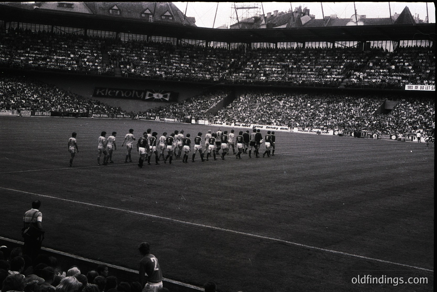 Black-and-white stadium shot of a pre-match football (soccer) team walkout, likely 1960s–1970s. Players in white kits line up on the pitch, flanked by spectators in tiered stands. Banner reads "The Derby."