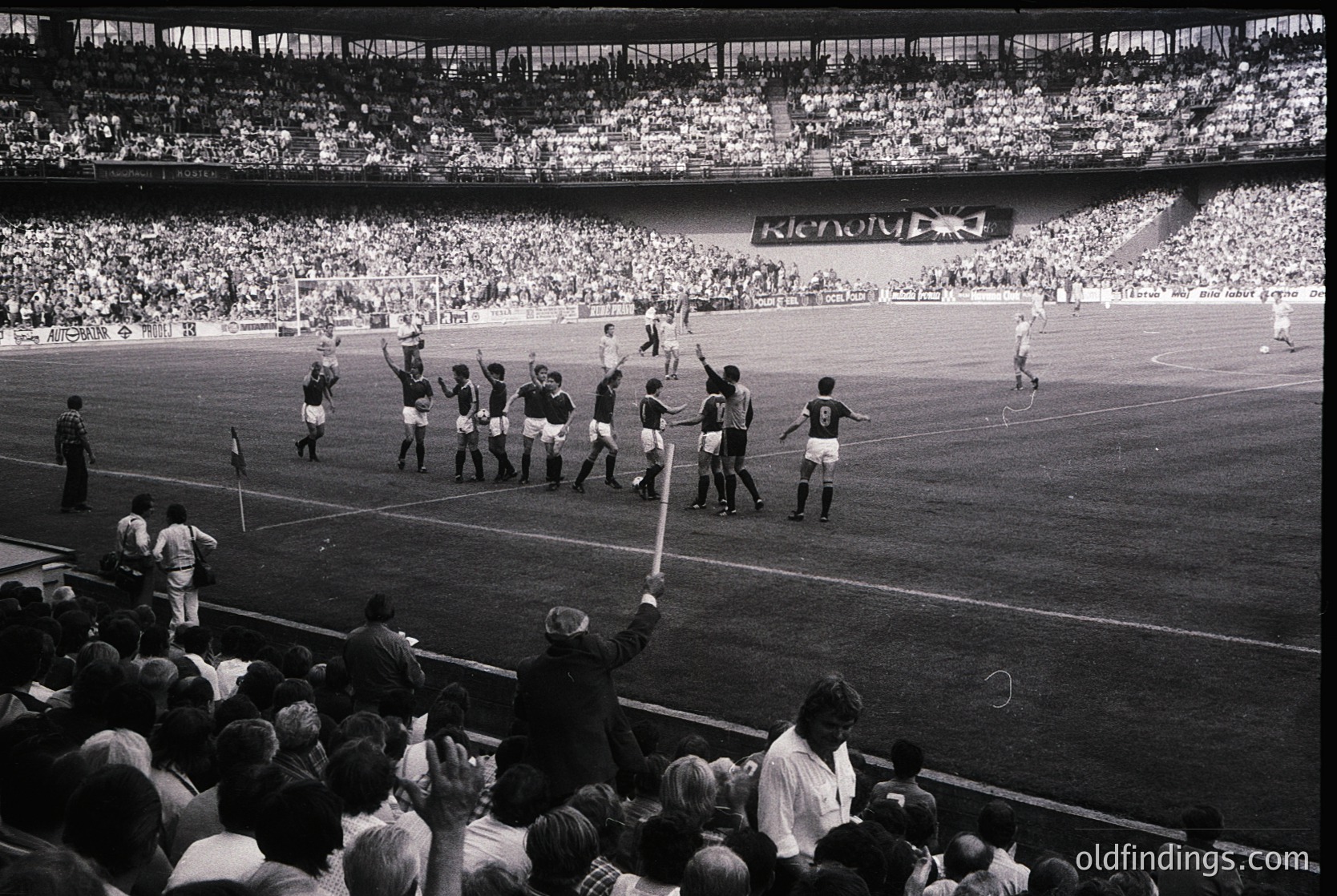 Black-and-white stadium shot of a post-match celebration, likely UEFA Champions League or European Cup era (1960s–1970s). Team in white jerseys raises arms in triumph, referee and officials present. Packed stands suggest high attendance, with "RICOH" banner visible. Iconic moment of European football history.