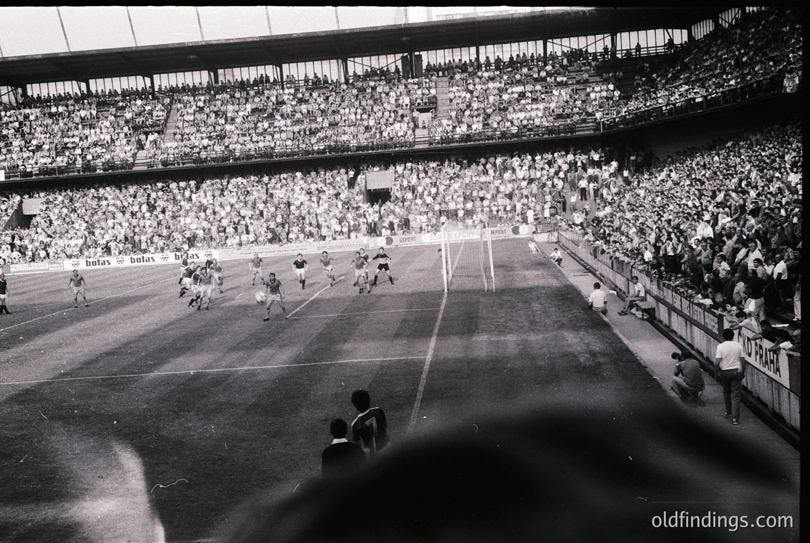 Vintage black-and-white stadium shot capturing a packed 1960s European football match. Players in white and dark jerseys contest near the goal, with a referee overseeing. Crowd fills tiered stands, creating a sea of spectators. Advertisements for "Bayer" and "Varta" visible on banners. Architectural details include curved roofing and tiered seating.