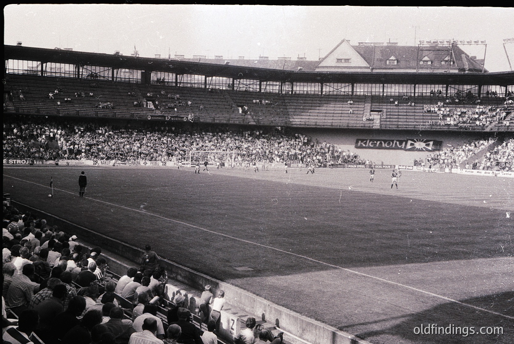 Vintage black-and-white stadium shot showing a packed 1960s-era football match. Multi-tiered stands filled with spectators, players in action on the field, and "Ketovimpex" banner visible. Architectural details include tiered seating and a central roof structure.