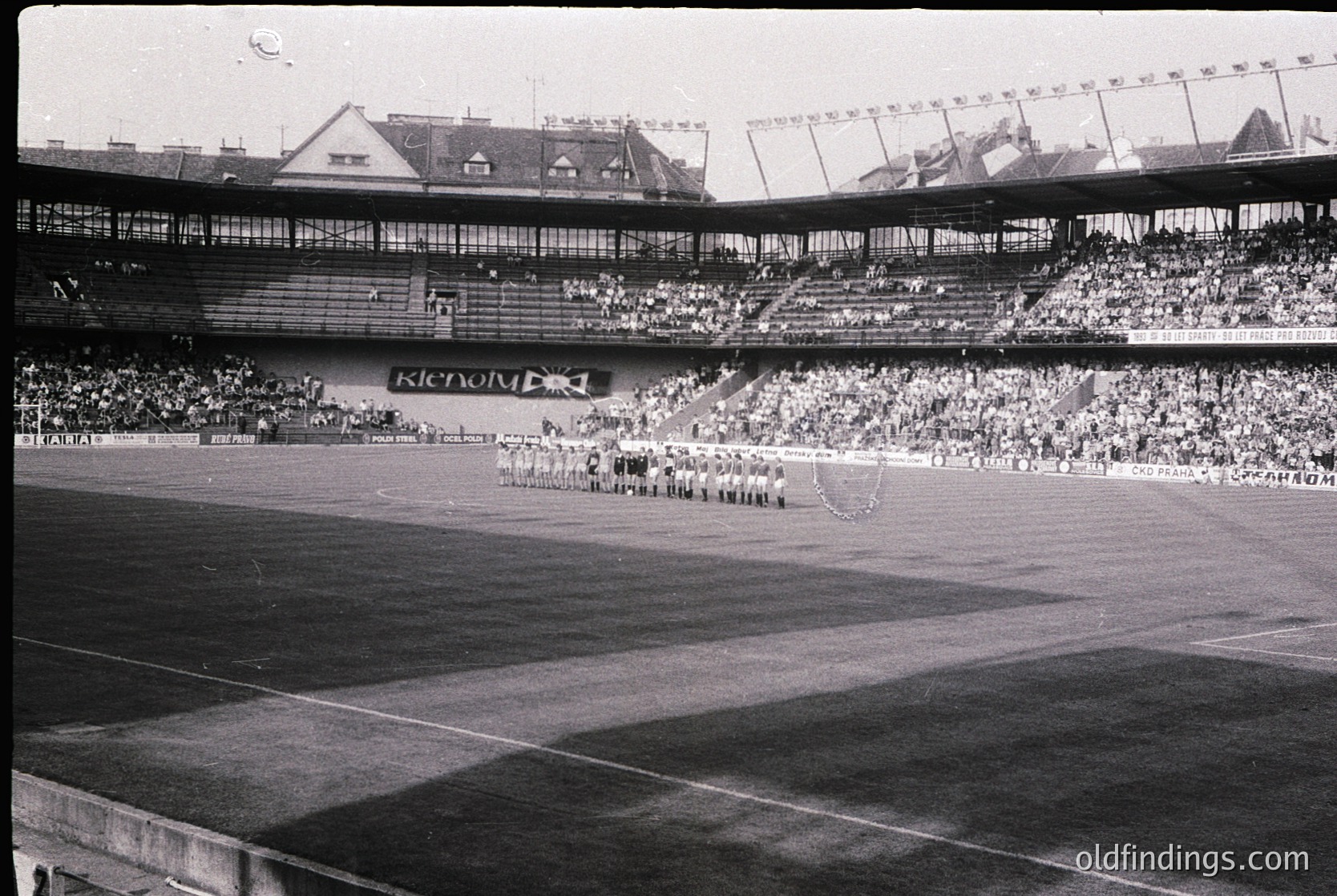 Black-and-white stadium shot featuring a large crowd in tiered seating, likely from the 1950s–1960s. Prominent "Kienzl" signage suggests a European venue. Uniformed athletes and officials stand on the field, possibly during an opening ceremony or pre-game event.