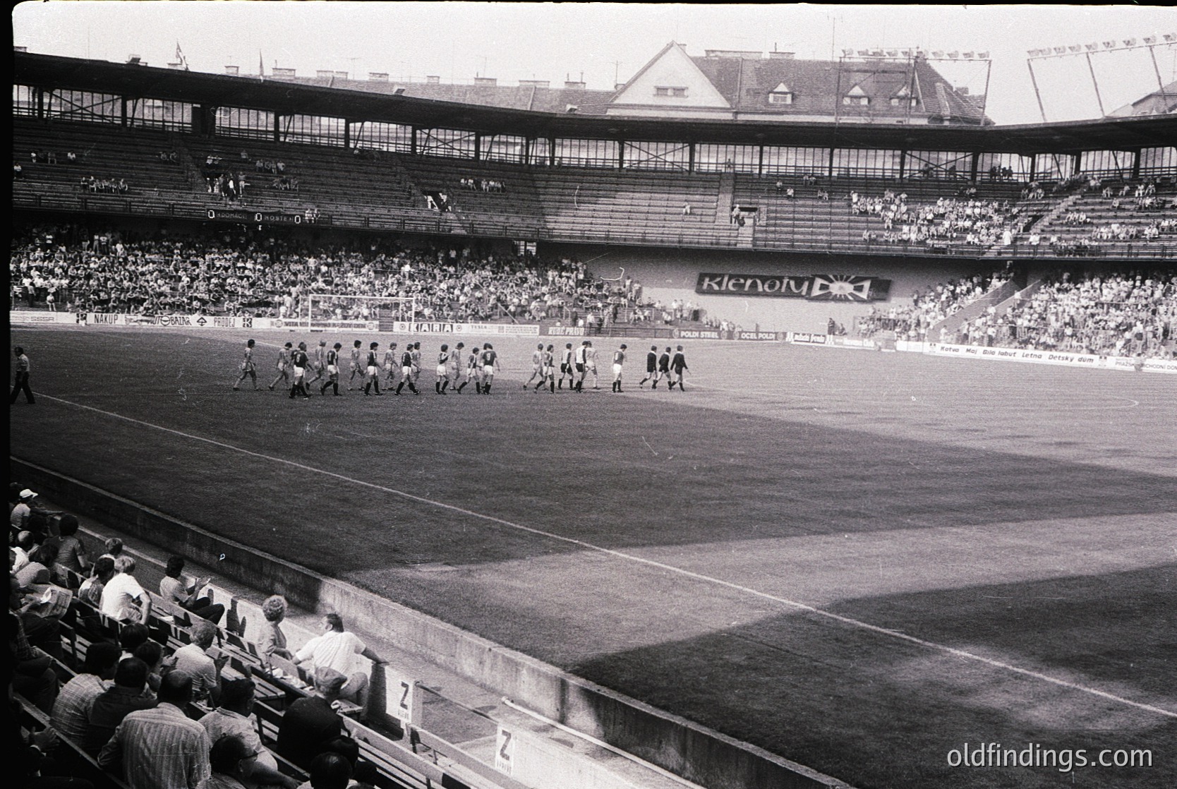 Black-and-white shot of a mid-20th-century stadium with players in formation, likely pre-game warm-up. Multi-tiered seating with sparse crowd, visible "Klenov" banner. Classic stadium architecture with covered stands and open-air lower sections.