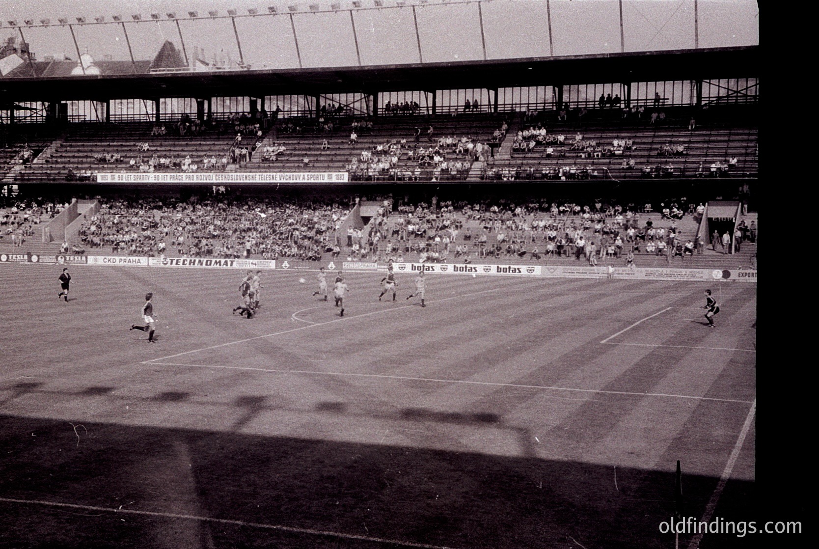 Vintage black-and-white stadium shot showing a packed 1960s-era football match. Players in striped jerseys engage in action on a grass pitch, with visible sponsor banners ("Steelkomat," "Bulgas," "Betas"). Crowd fills tiered stands, indicating high attendance. Architectural details include concrete seating and exposed roof trusses.