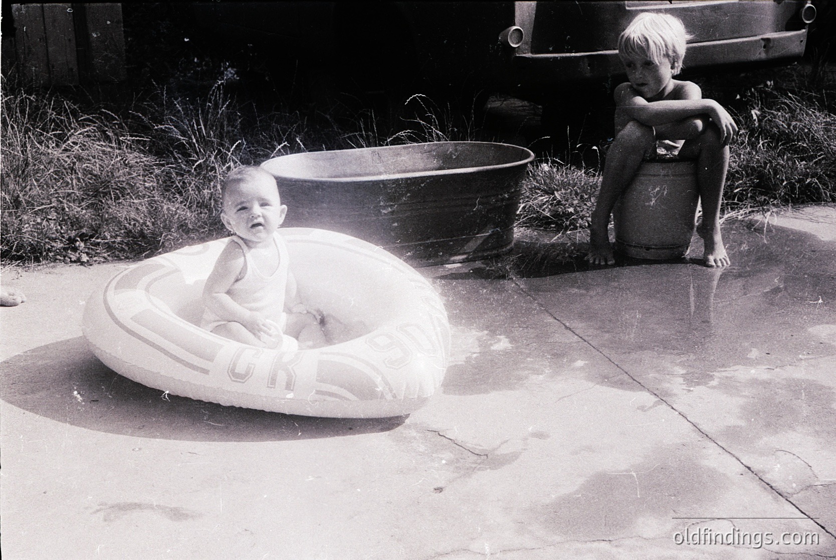 Mid-20th century backyard scene: infant in a white inner-tube on concrete, older child seated on a barrel. Classic 1950s–60s suburban playtime.