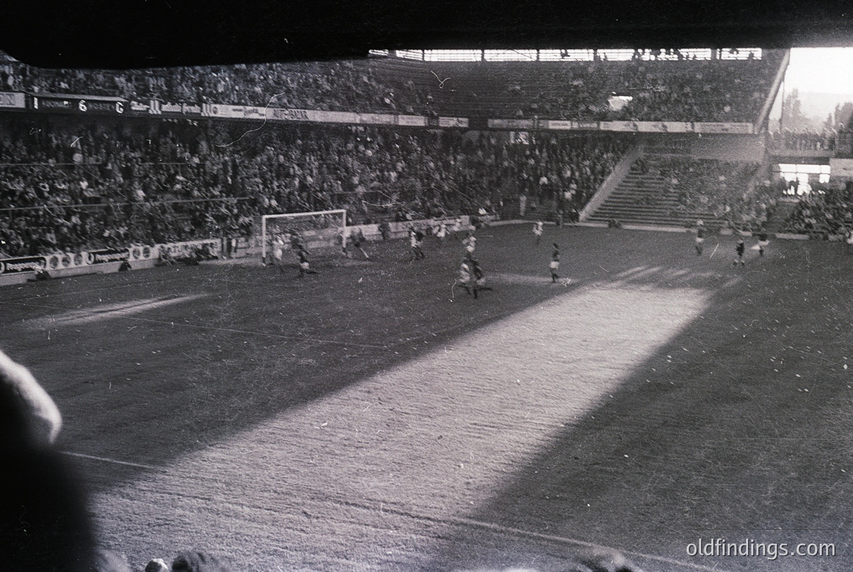 Black-and-white stadium shot showing a packed arena with players on a muddy pitch, likely a 1960s–70s European football match. Crowds fill tiered stands, with visible advertising boards and floodlights illuminating the field. Atmosphere captures mid-action, possibly a penalty kick or corner.