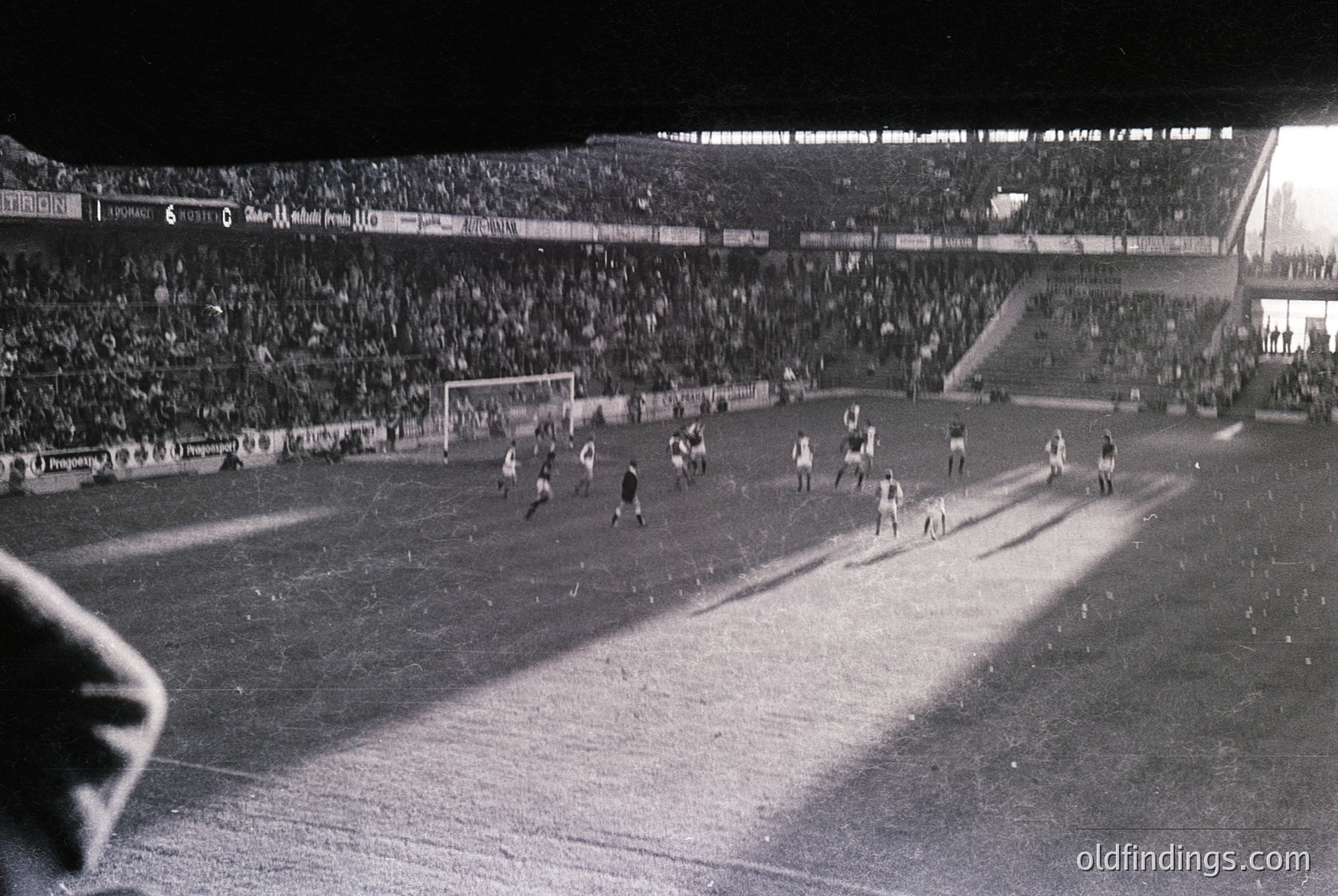 Black-and-white shot of a packed stadium during a 1960s–1970s football match, likely in Europe. Crowds fill tiered stands under floodlights, with players in white and dark uniforms engaged in action near the goal. Advertising banners (e.g., "CIGA") and stadium signage visible.
