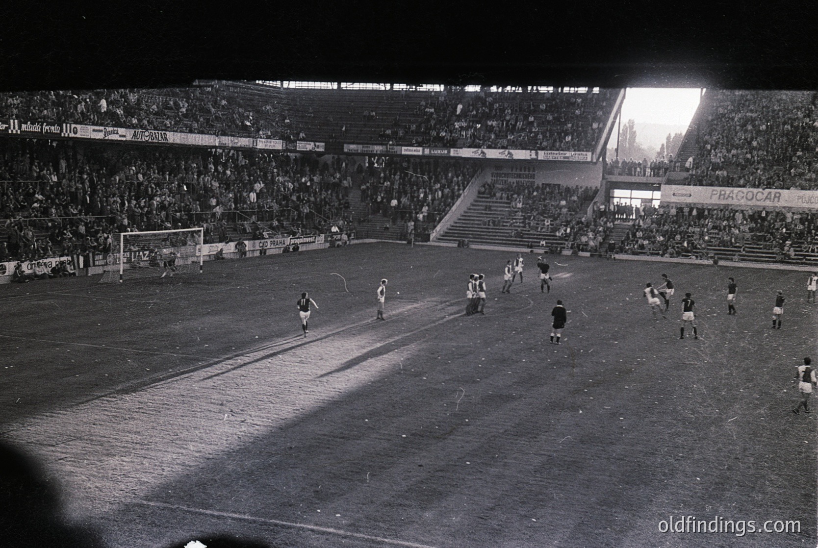 Black-and-white nighttime soccer match in a large stadium, featuring a packed crowd under floodlights. Players in dark uniforms chase the ball on a muddy pitch. Advertisements like "FRAGOCAR" and "BANCO" visible on stadium boards. Atmosphere reflects mid-20th century European football culture.