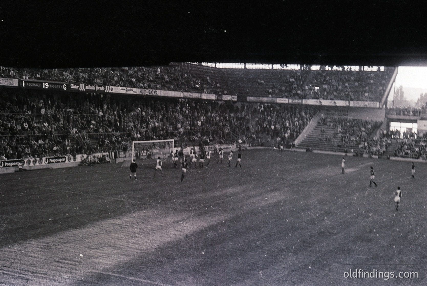 Black-and-white shot of a packed stadium during a nighttime soccer match, featuring a packed stadium with floodlit field and goal. Players in vintage uniforms engage in action near the goalpost, with spectators densely filling tiered stands. Advertising boards display text in Spanish, suggesting a European league setting. Likely 1960s–1970s era.