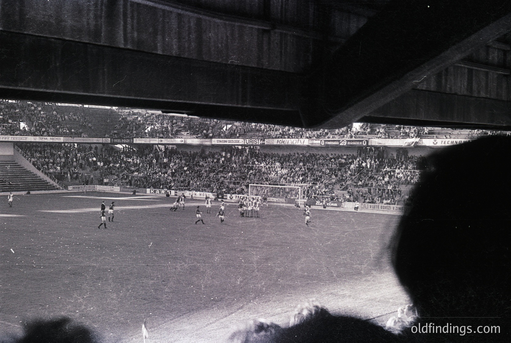Black-and-white shot of a packed stadium during a mid-20th-century football match. Crowds fill tiered stands, with players and ball visible on the field. Architectural details include curved roofing and floodlights. Likely European setting, 1950s–1960s era.