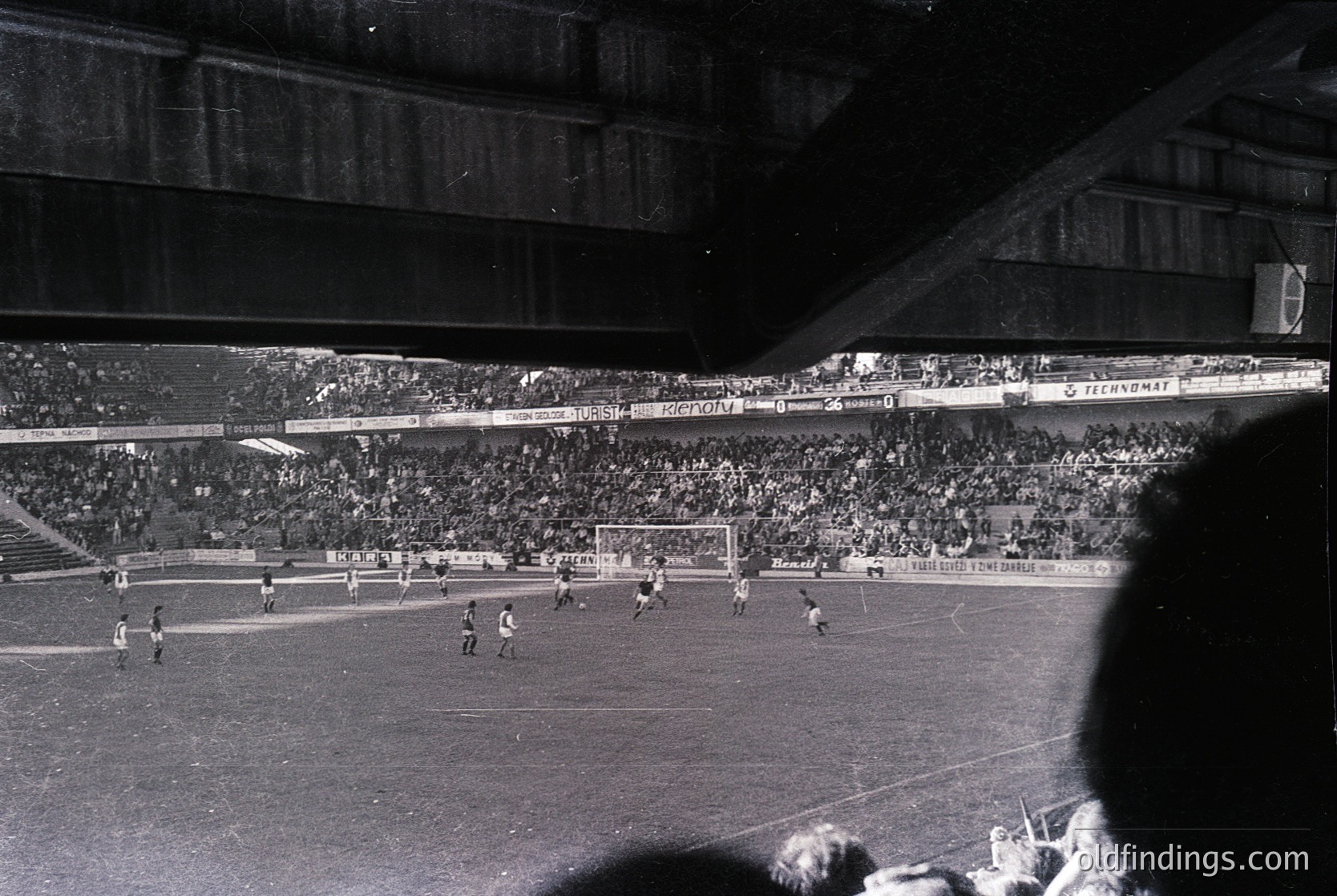 Black-and-white stadium shot capturing a mid-20th-century football match. Crowded stands with sparse advertising (e.g., "Technomat") and a packed pitch featuring players in classic striped jerseys. Architectural details include tiered seating and floodlights. Likely European, 1950s–1960s.