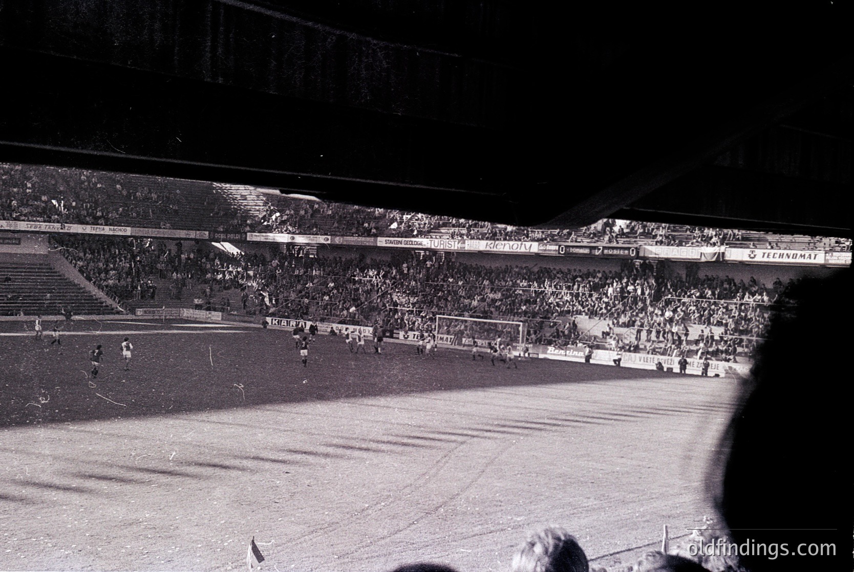 Vintage black-and-white shot of a packed stadium during a soccer match, likely 1960s–1970s. Crowds fill tiered stands under floodlights, with visible advertisements like "Tecnomat" and "Proprietà." Snow covers the field, indicating winter play. Architectural details include curved seating and a central floodlight structure.