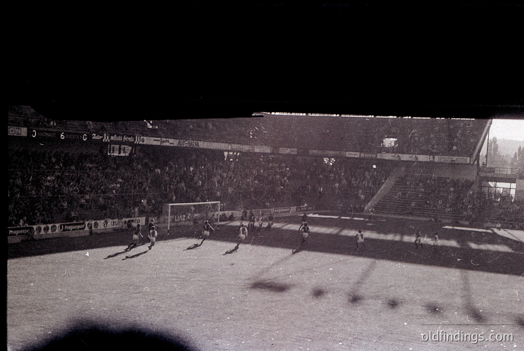 Vintage black-and-white shot of a packed stadium during a nighttime football match. Crowds fill tiered stands under floodlights, with visible banners advertising brands like "Laz" and "Republika." Players in classic striped jerseys and shorts engage in action on the pitch. Architectural details include a multi-level stadium with open upper sections. Likely Eastern European, 1960s–1980s era.