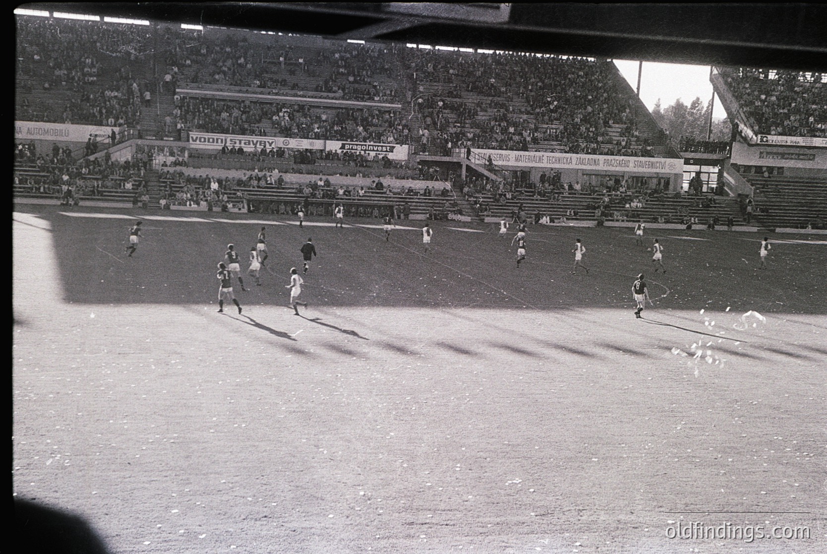 Black-and-white shot of a packed stadium during a mid-20th century football match. Visible banners advertise brands like "Vooni" and "Staviv," with Cyrillic text suggesting Eastern European location. Players in vintage striped jerseys and shorts engage in action on a muddy field. Crowd fills tiered stands, indicating high attendance.