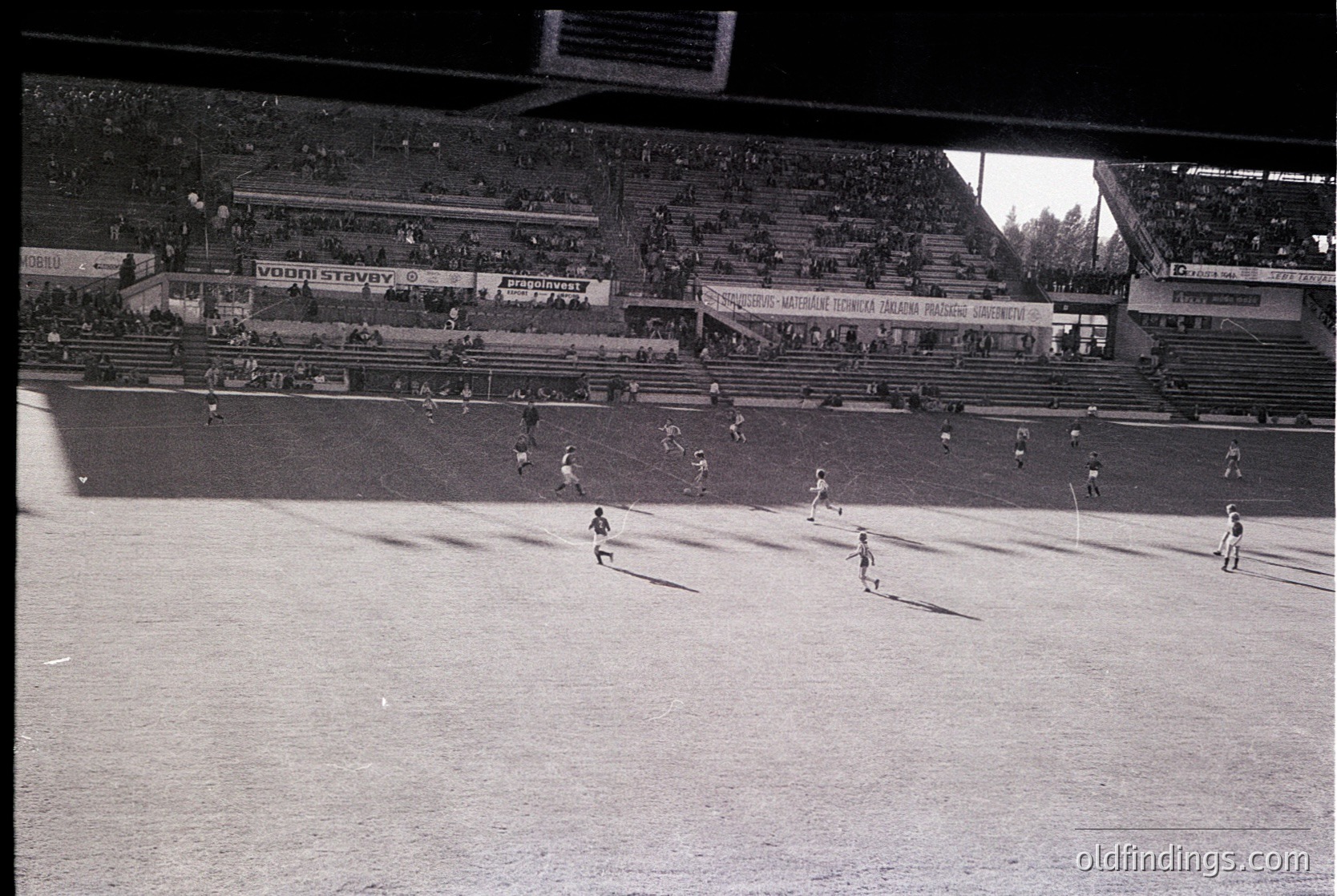 Vintage black-and-white shot of a packed stadium during a soccer match, featuring midfield action with players in motion. Advertisements like "Vodni stavby" and "Sokol" visible on stands, indicating Eastern European setting. Crowd fills tiered seating, suggesting 1960s–1970s era.