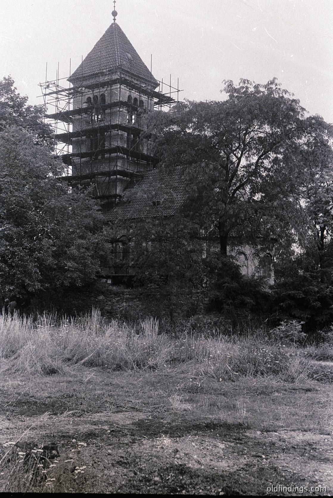Historic stone tower under restoration, featuring a conical roof and multi-tiered balcony structure. Scaffolding surrounds the upper levels, indicating mid-20th century preservation efforts. Surrounded by dense foliage and overgrown grass, suggesting a rural or park setting.