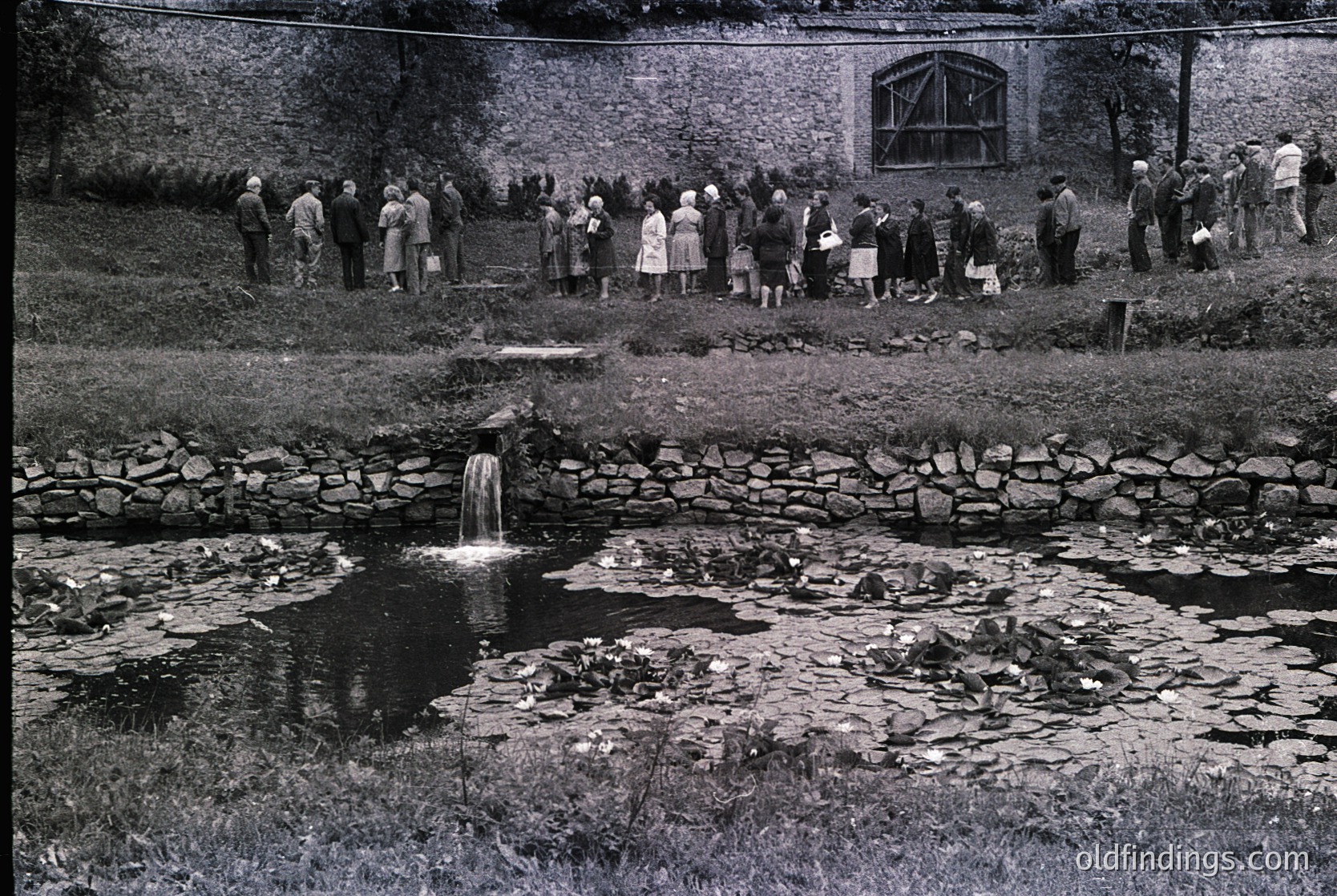 Mid-20th century gathering by a rustic stone fountain in a rural setting. Crowd of ~40 people in 1950s-60s attire—men in suits, women in dresses—standing on grassy bank. Stone wall and wooden structure with arched windows in background. Fountain spills into shallow, moss-covered pool.