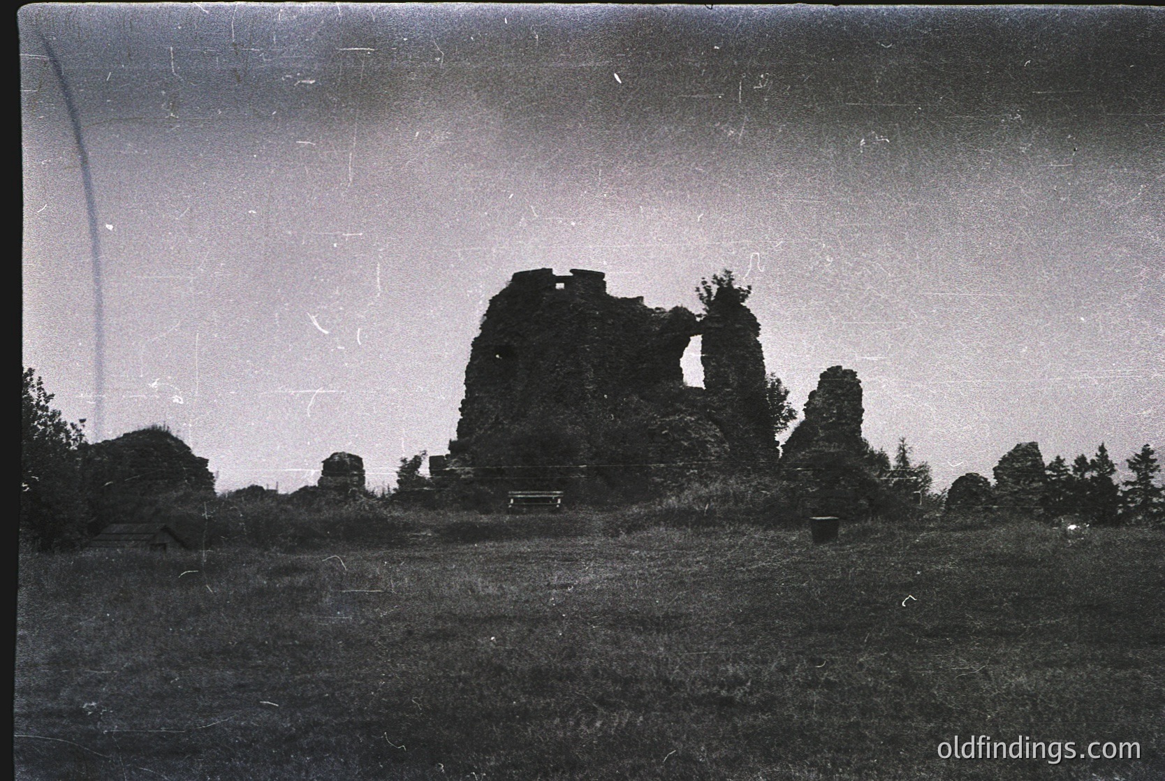 Vintage black-and-white photo of a ruined stone tower with crumbling walls and overgrown vegetation, set in a grassy field. Silhouetted trees frame the scene, suggesting a historic or abandoned site. Likely early-to-mid 20th century based on grain and contrast.