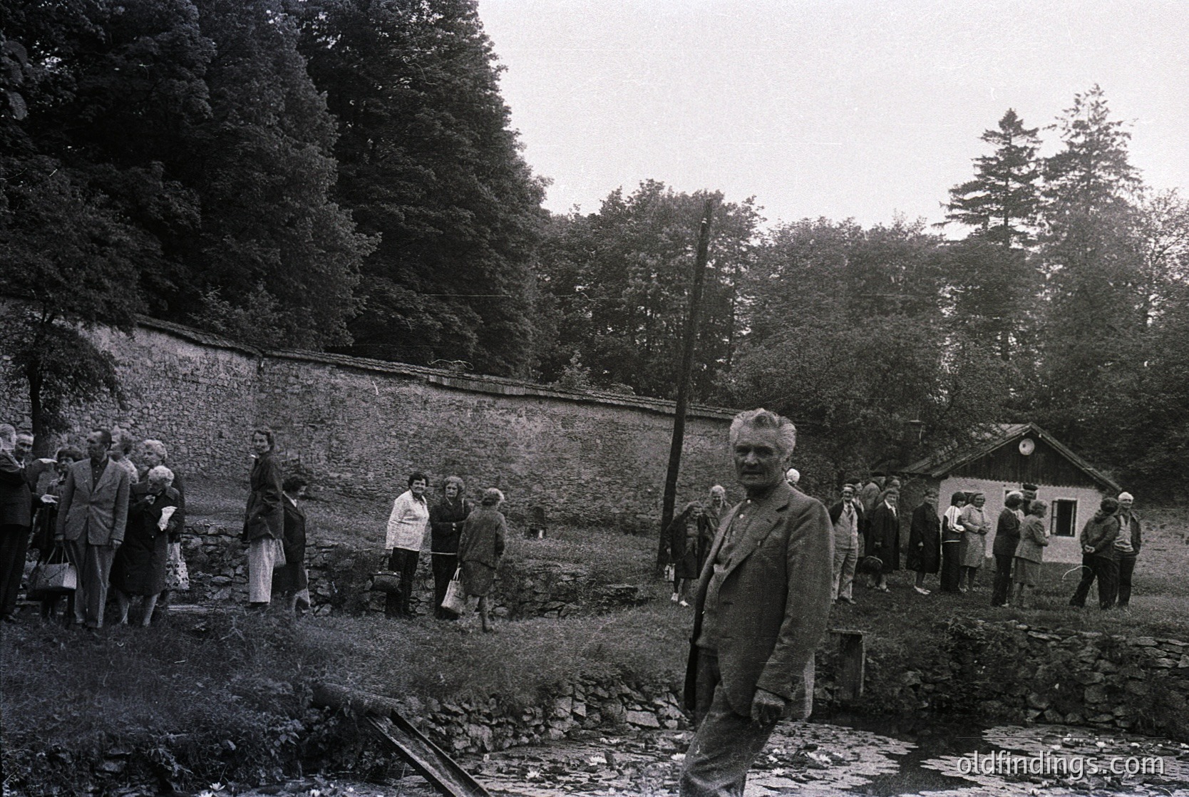 Mid-20th century black-and-white photo of a rural gathering near a stone wall and small wooden structure. Central figure in suit poses near a shallow water area, surrounded by ~15 people in period attire. Dense forest and overcast sky frame the scene.