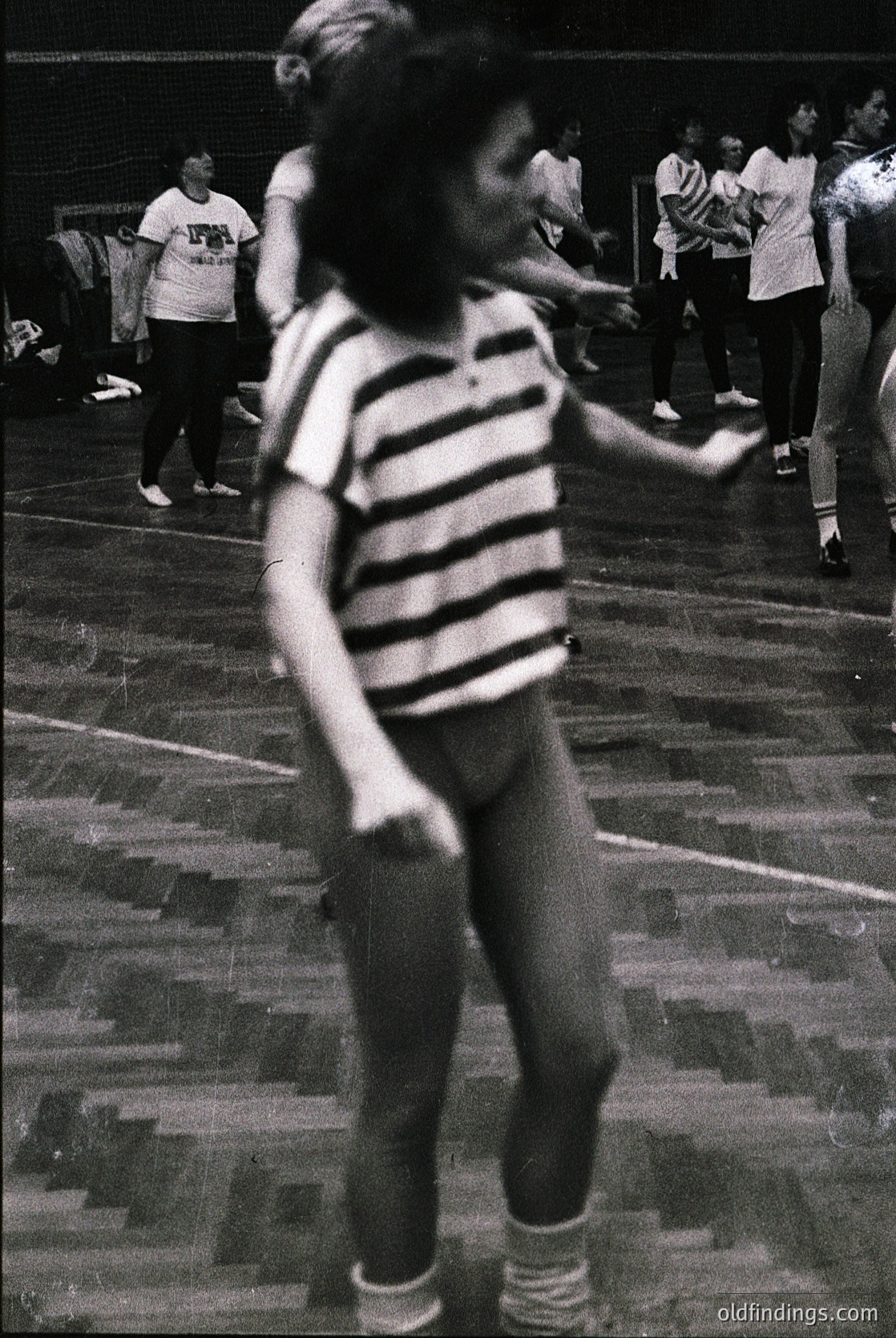 Vintage indoor gym scene with blurred motion—child in striped shirt and shorts mid-jump, wooden floor, blurred spectators in white shirts. Likely 1960s–1970s physical education or sports event.