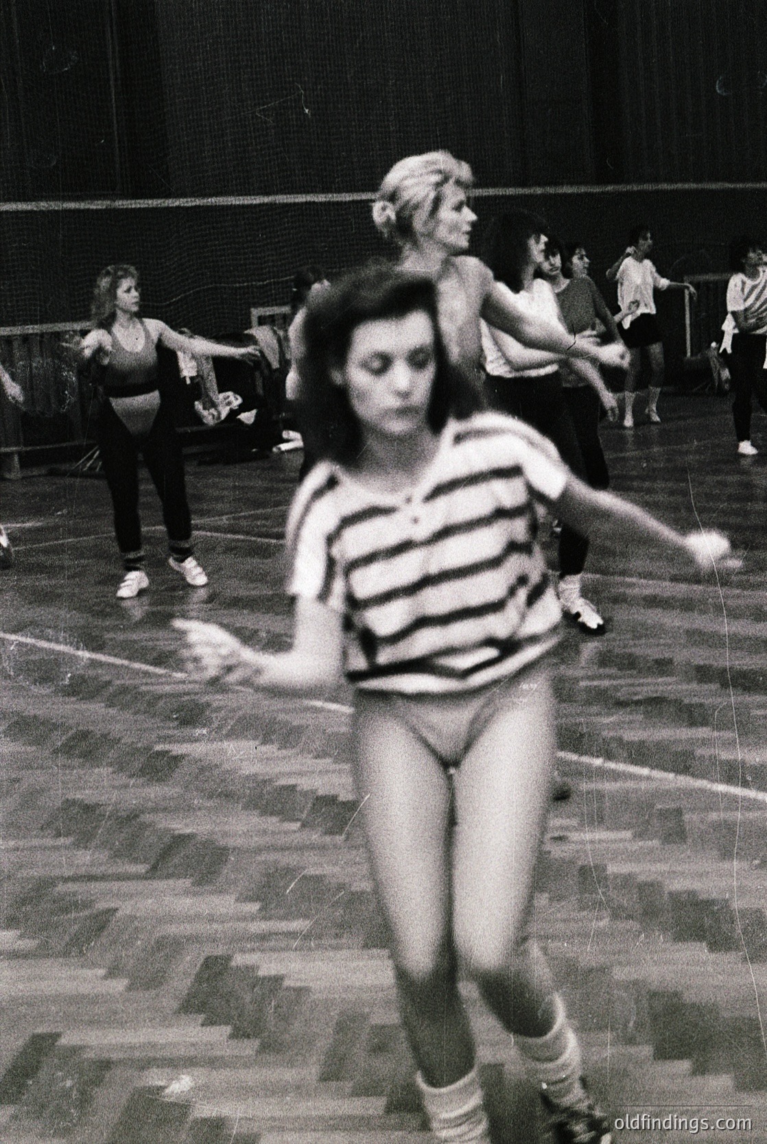 Vintage black-and-white gym class scene, 1960s-70s. Women in form-fitting leotards and knee-high socks practice dance or gymnastics on a polished wooden floor. Dynamic pose suggests rhythmic movement, likely aerobic or modern dance. Indoor setting with blurred background indicating motion.