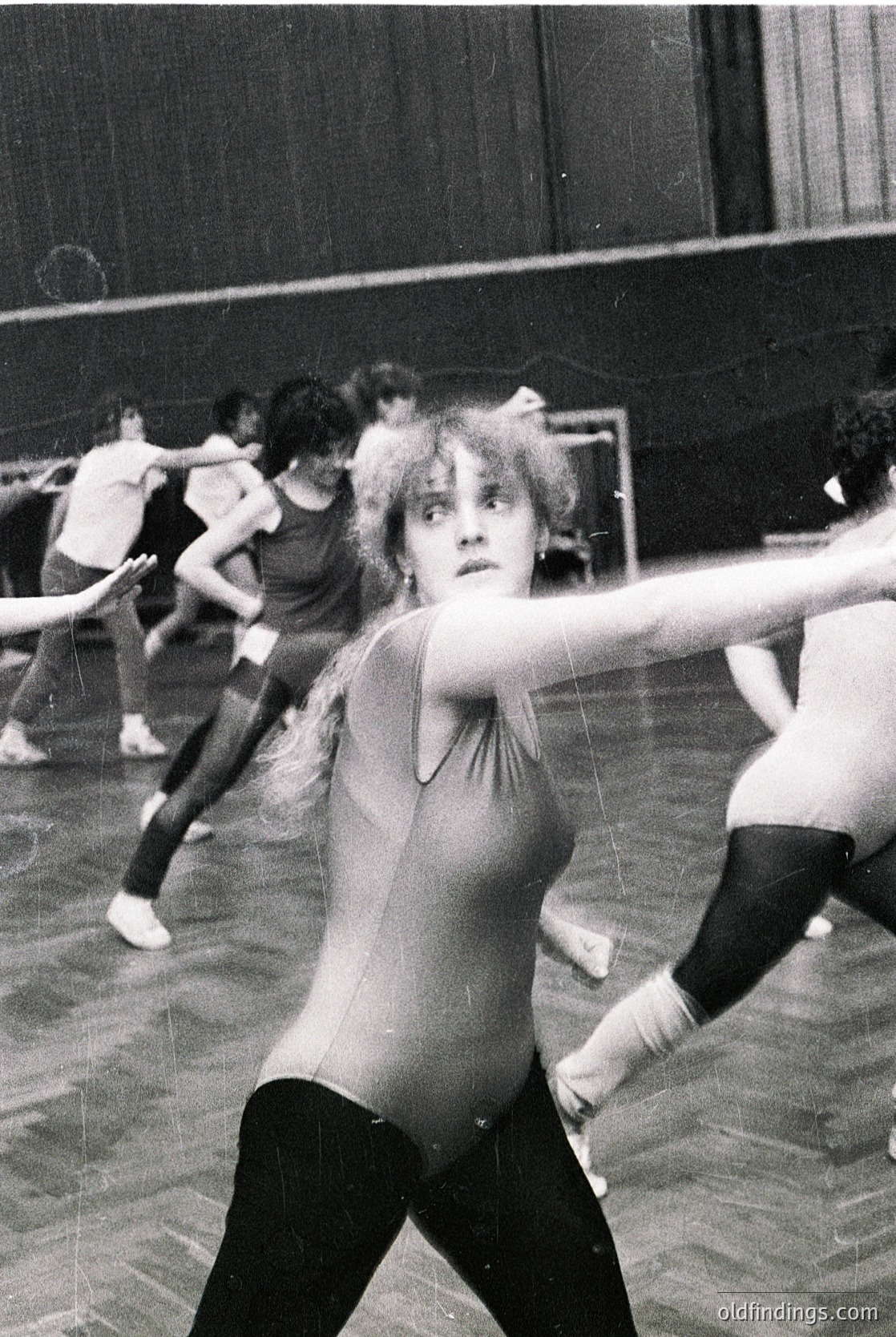 Black-and-white shot of a dance studio session, likely 1960s–1980s. Foreground dancer in sleeveless top and tights, mid-movement with arms extended. Indoor setting with industrial lighting and blurred peers practicing.