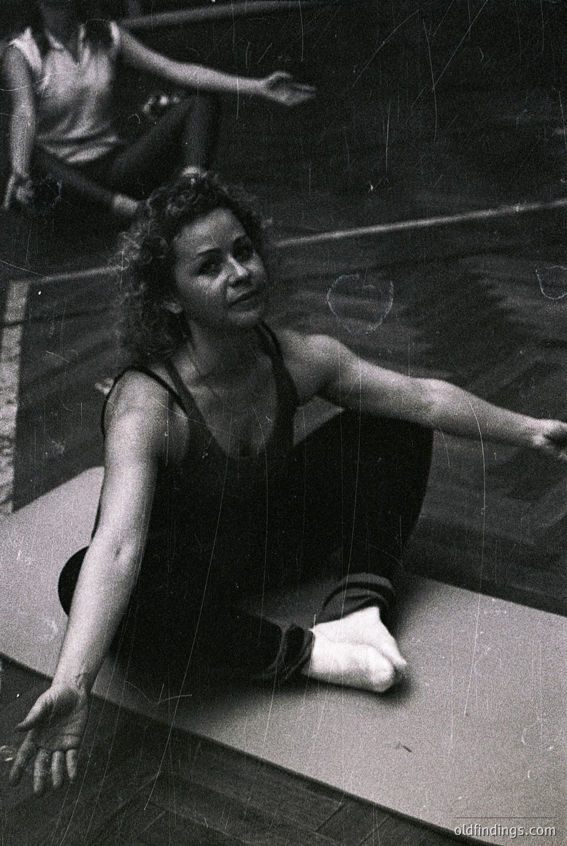 Vintage black-and-white shot of a gymnast in mid-dynamic pose, likely mid-20th century. The woman’s sleek leotard and extended limbs suggest training or rehearsal. Reflections on the mat hint at studio lighting.