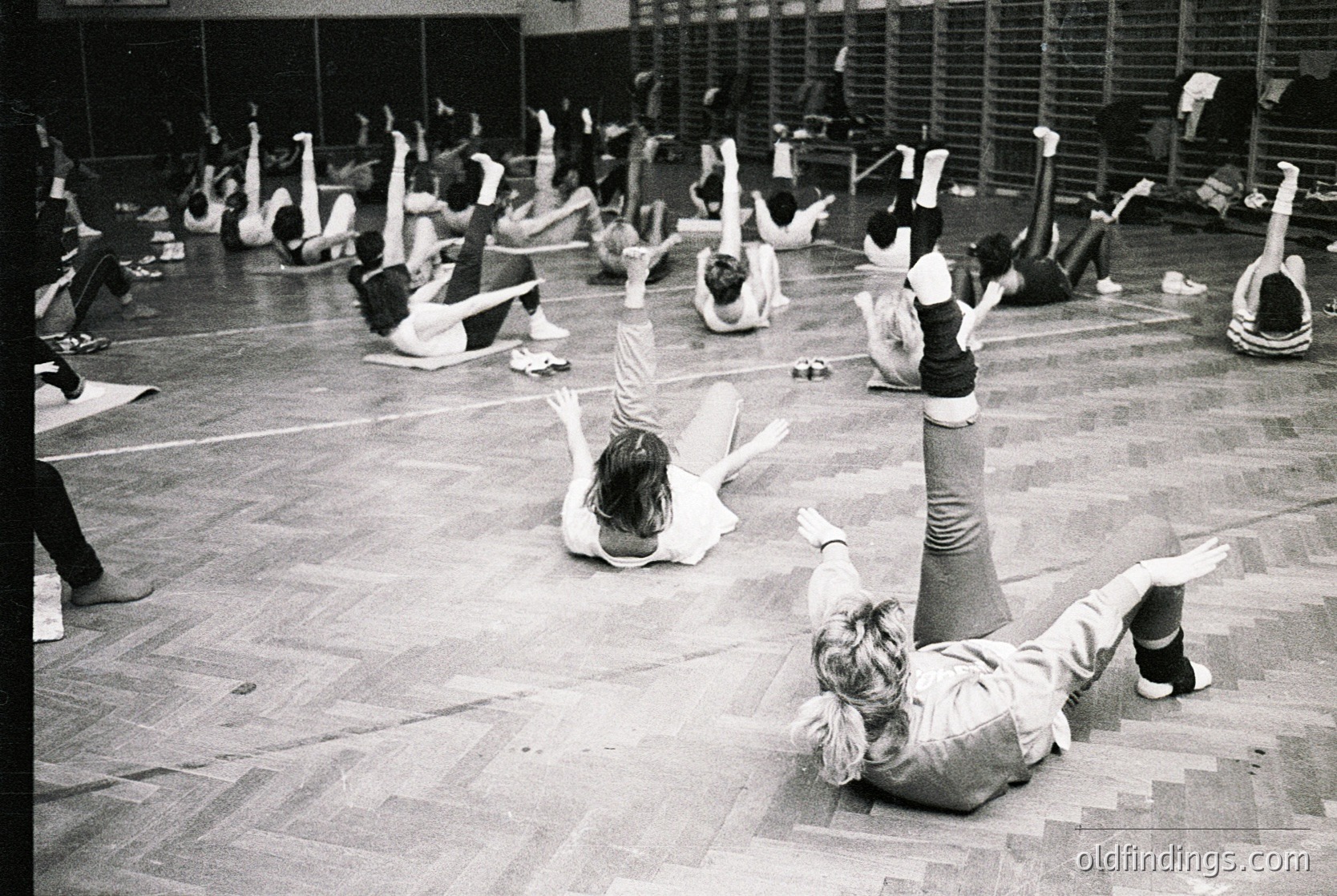 Group aerobics class in an indoor gym, likely 1970s–1980s. Participants perform floor exercises in unison, wearing leotards and leg warmers. Wooden bleachers line the perimeter, and a brick wall with windows provides natural light.