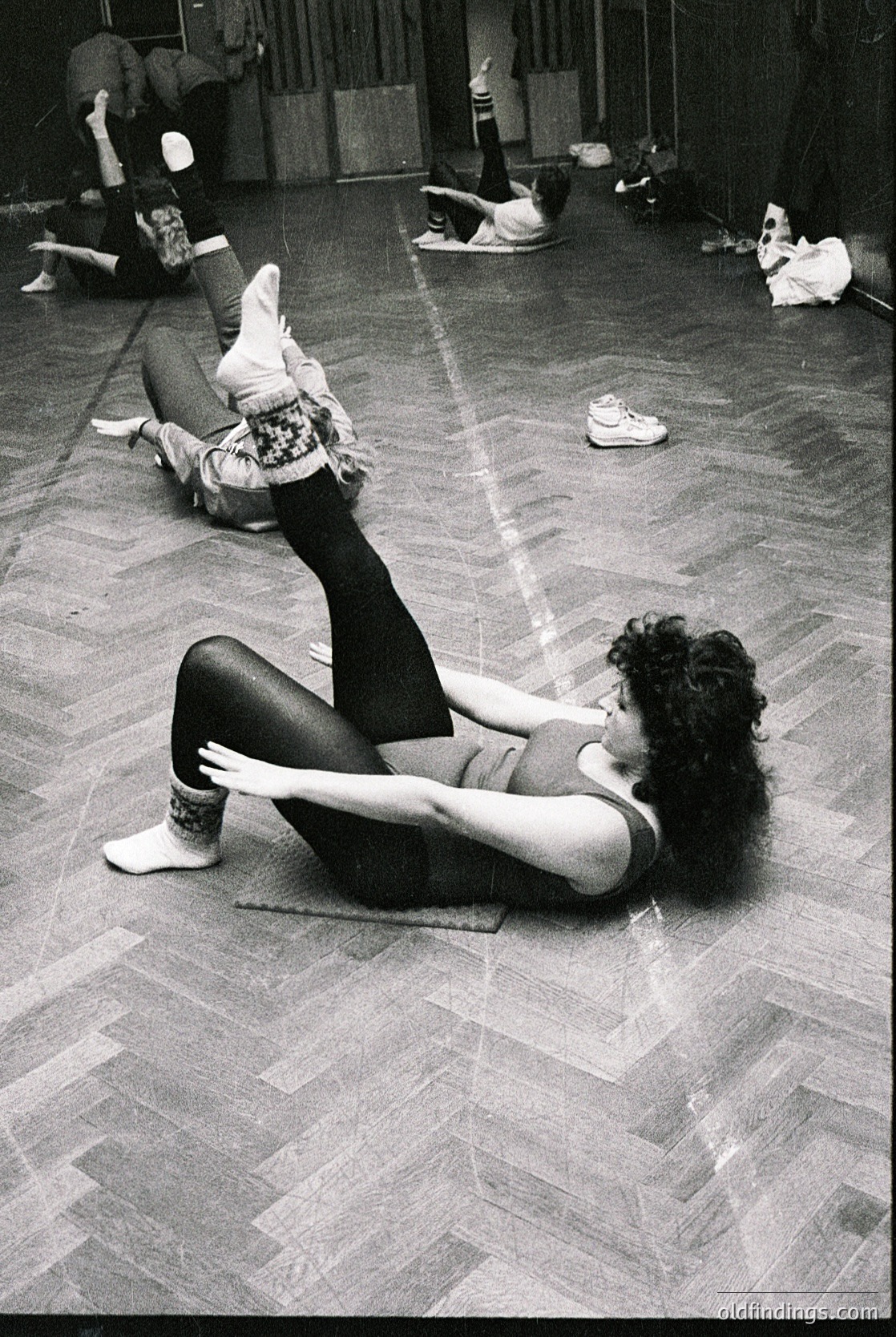 Black-and-white studio shot of dancers in mid-rehearsal, 1960s–1970s. Two women stretch on wooden floor—one seated with legs extended, the other kneeling with bent knees. Minimalist ballet attire (leotards, tights, ballet slippers) and scattered props (barre, shoes) suggest a professional dance studio.