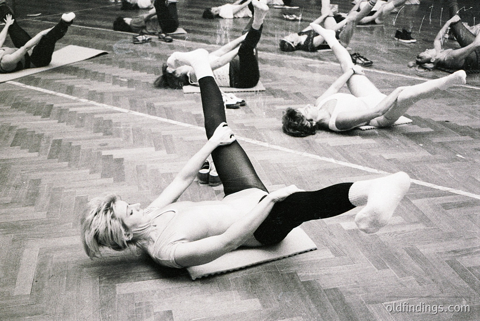 Group fitness class in a gymnasium, 1970s. Women perform floor exercises on mats in a large, wood-floored hall. Foreground subject demonstrates leg lifts while others stretch or balance. Minimalist, functional interior with parallel lines of mats.