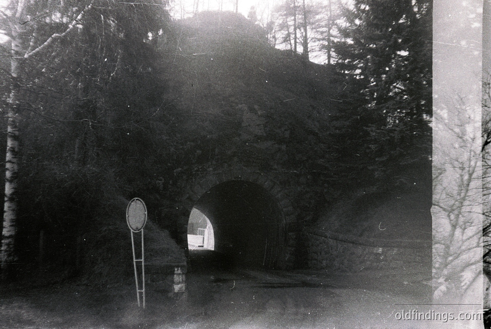 Vintage black-and-white tunnel entrance with directional road sign indicating a right turn. Surrounded by dense forest and rocky terrain, suggesting a rural or mountainous region. Likely early-to-mid 20th century based on photographic style and sign design.