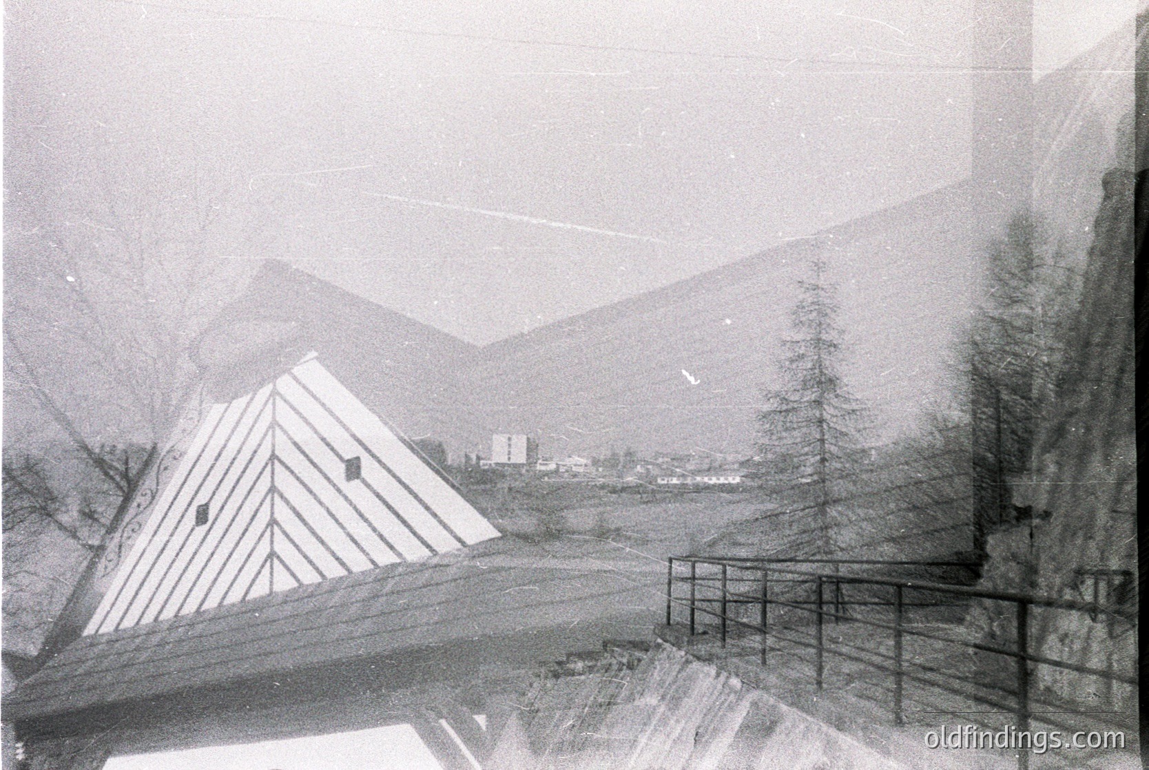 Vintage alpine lodge framed by snow-laden pine trees, set against misty mountain peaks. Geometric, pyramid-roofed structure with small windows, likely mid-20th century. Rustic wooden railings and rocky terrain suggest a remote, high-altitude location.