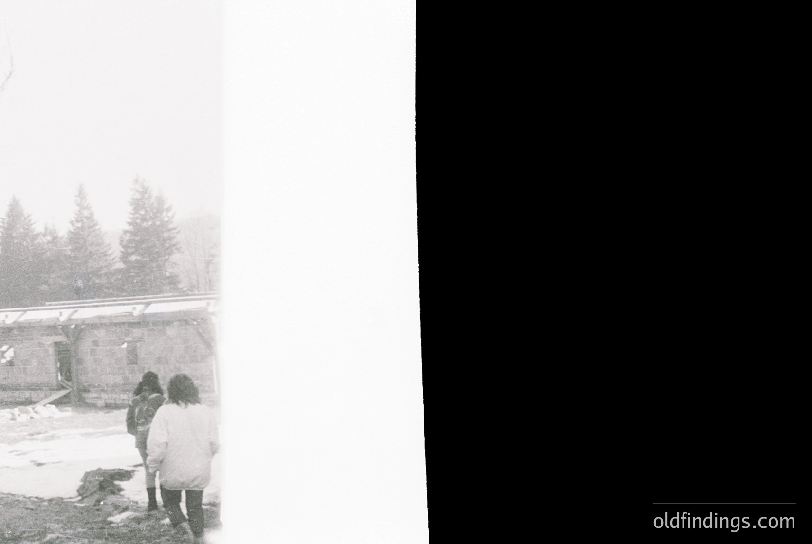 Black-and-white winter scene featuring three figures in heavy coats near a snow-covered structure. Dense fog obscures distant coniferous trees, creating a moody, atmospheric backdrop. Likely mid-20th century due to clothing and film grain. Ideal for vintage travel or historical research.