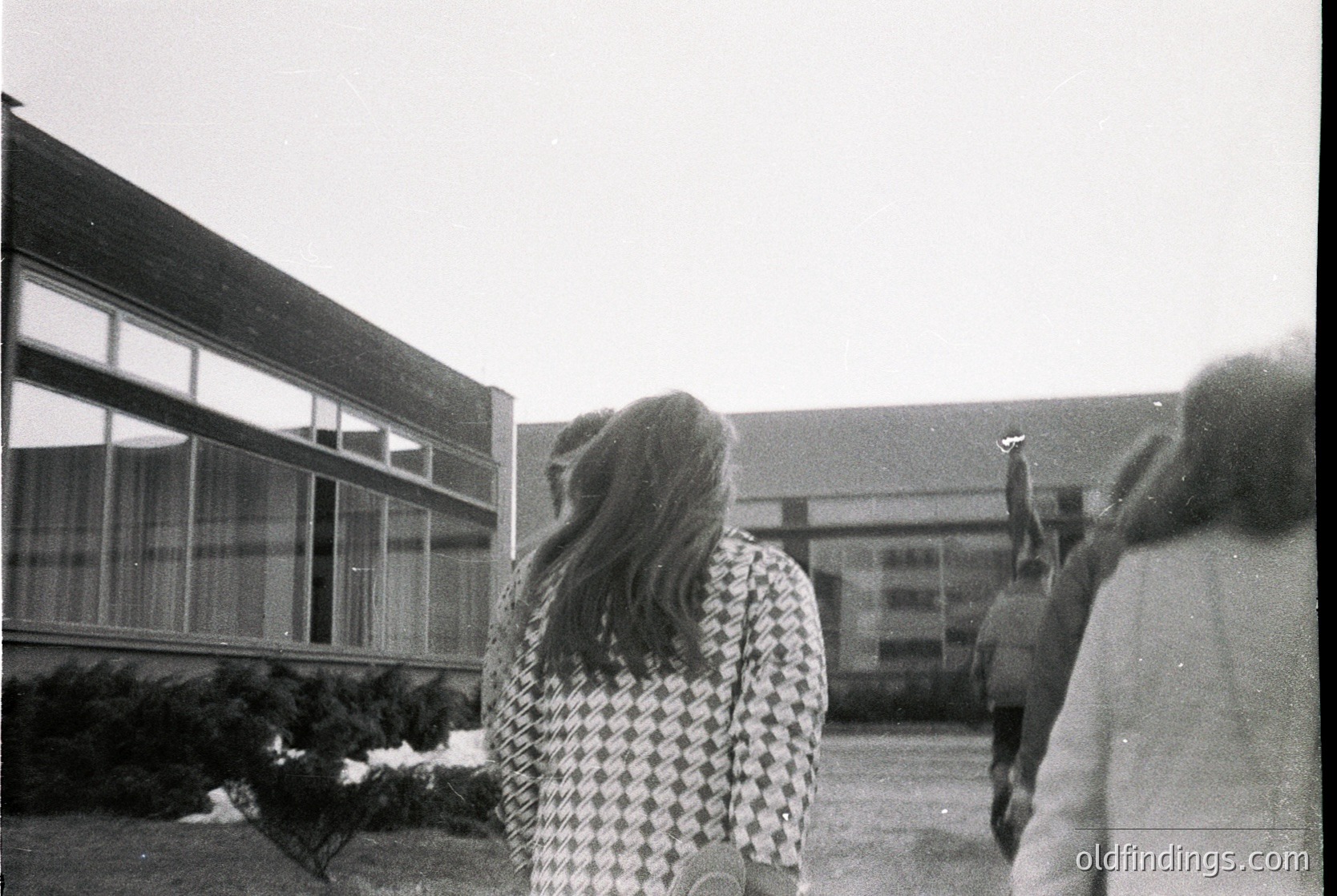 Black-and-white snapshot of two figures in mid-motion outside a mid-century modern building with large glass panels. One person wears a checkered blouse; the other, a light sweater. Snow patches on ground suggest winter. Likely 1960s–1970s, urban/suburban setting.
