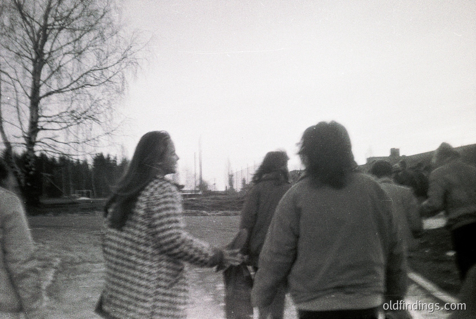 Black-and-white candid of three figures walking outdoors, holding hands. The woman in striped sweater leads, with blurred urban/suburban background showing trees, bare branches, and distant buildings. Mid-20th century streetwear suggests or . Potential study of social dynamics or urban life.